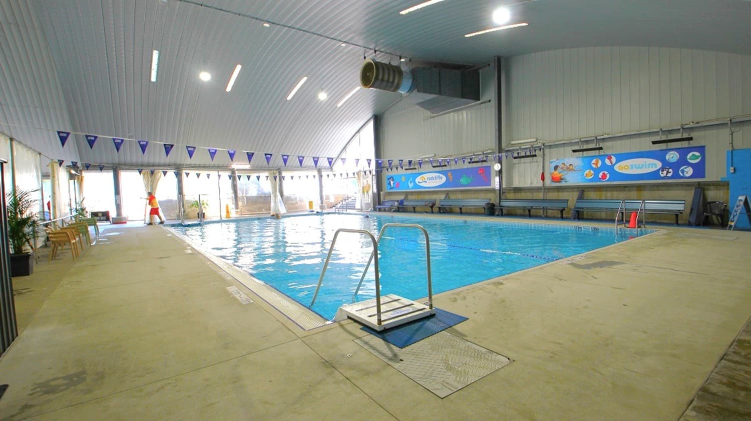 An indoor swimming pool with clear blue water is surrounded by a beige tiled deck. On the left, there are chairs and a few potted plants near large windows with curtains. A lifeguard stands by the poolside. The right side features colourful wall murals and benches. Overhead, the ceiling is high with bright lights and a large ventilation duct. Triangular flags are strung across the pool.