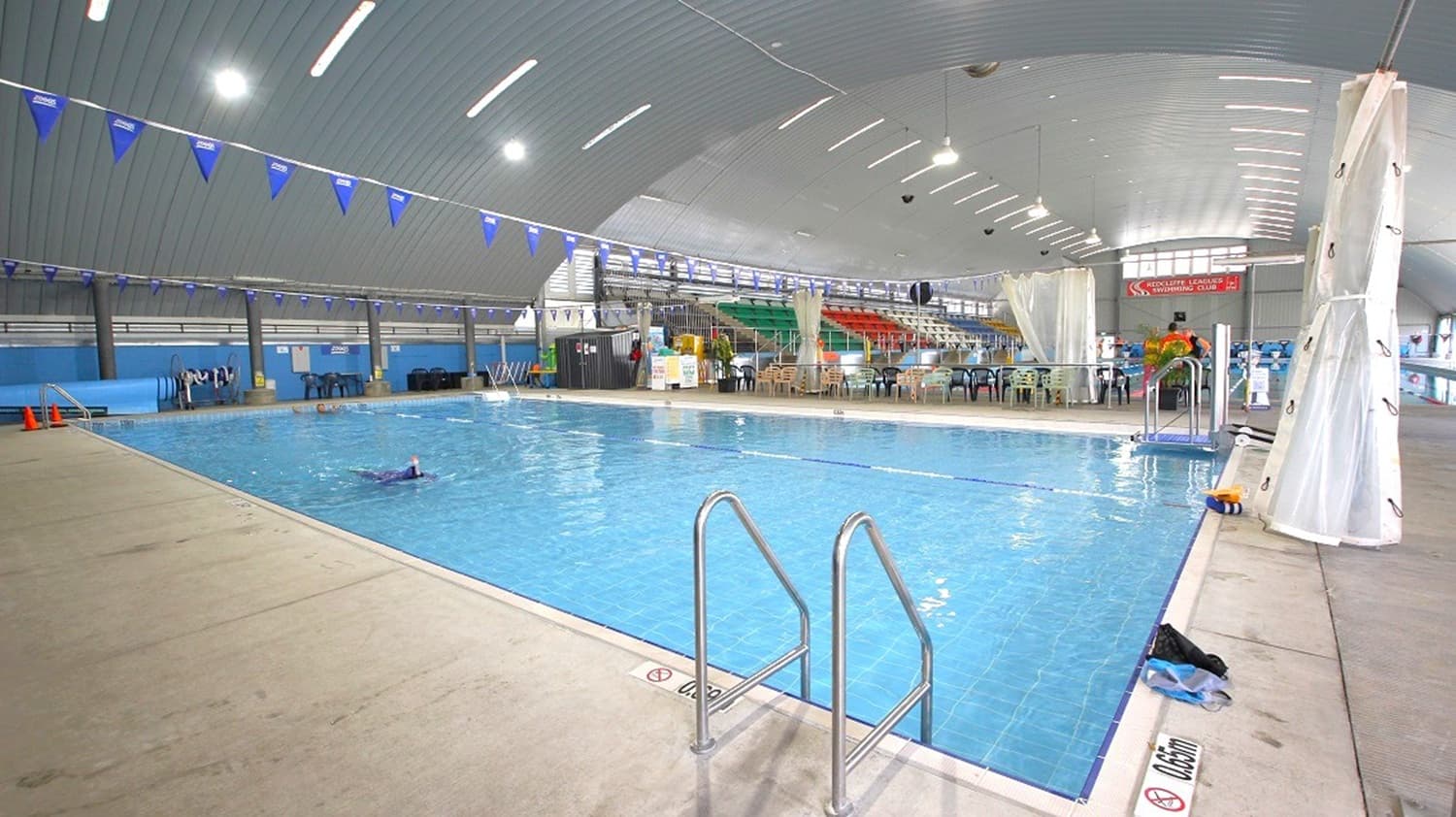 Indoor swimming pool with clear blue water, surrounded by a beige tiled deck. A metal ladder is in the foreground. Blue triangular flags hang above the pool. On the right, there are white curtains and a seating area with tables and chairs. The background features tiered seating with colourful chairs. The ceiling is arched with bright lights, and a swimmer is visible in the water.