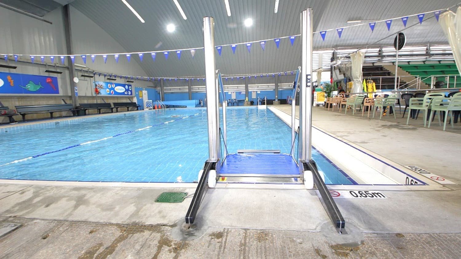 Indoor swimming pool with a metal accessibility lift in the foreground, leading into the water. The pool is surrounded by a tiled deck, with a depth marking of 0.65m visible. On the right, there are plastic chairs and tables, and a staircase leading to an upper level. The ceiling is high with bright lights, and blue triangular flags are strung across the pool. The left side features benches and colourful wall decorations.