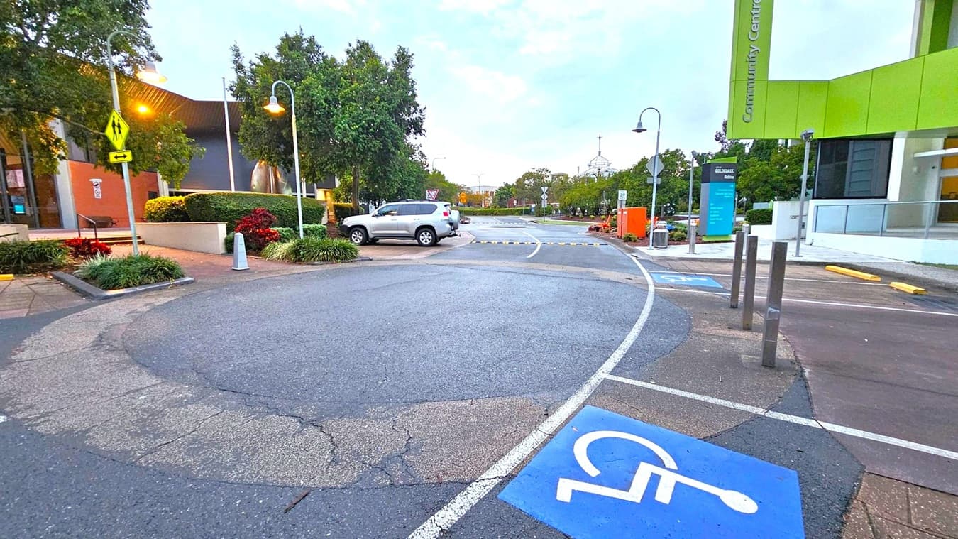 A street scene with a focus on a parking area. In the foreground, a blue disabled parking space is marked on the asphalt. On the left, there are trees and a building with a brown and grey facade. A silver SUV is parked nearby. On the right, a modern building with a green sign reading "Community Centre" is visible. Street lamps line the road, and there are various signs and bollards. The background features more trees and a clear sky.