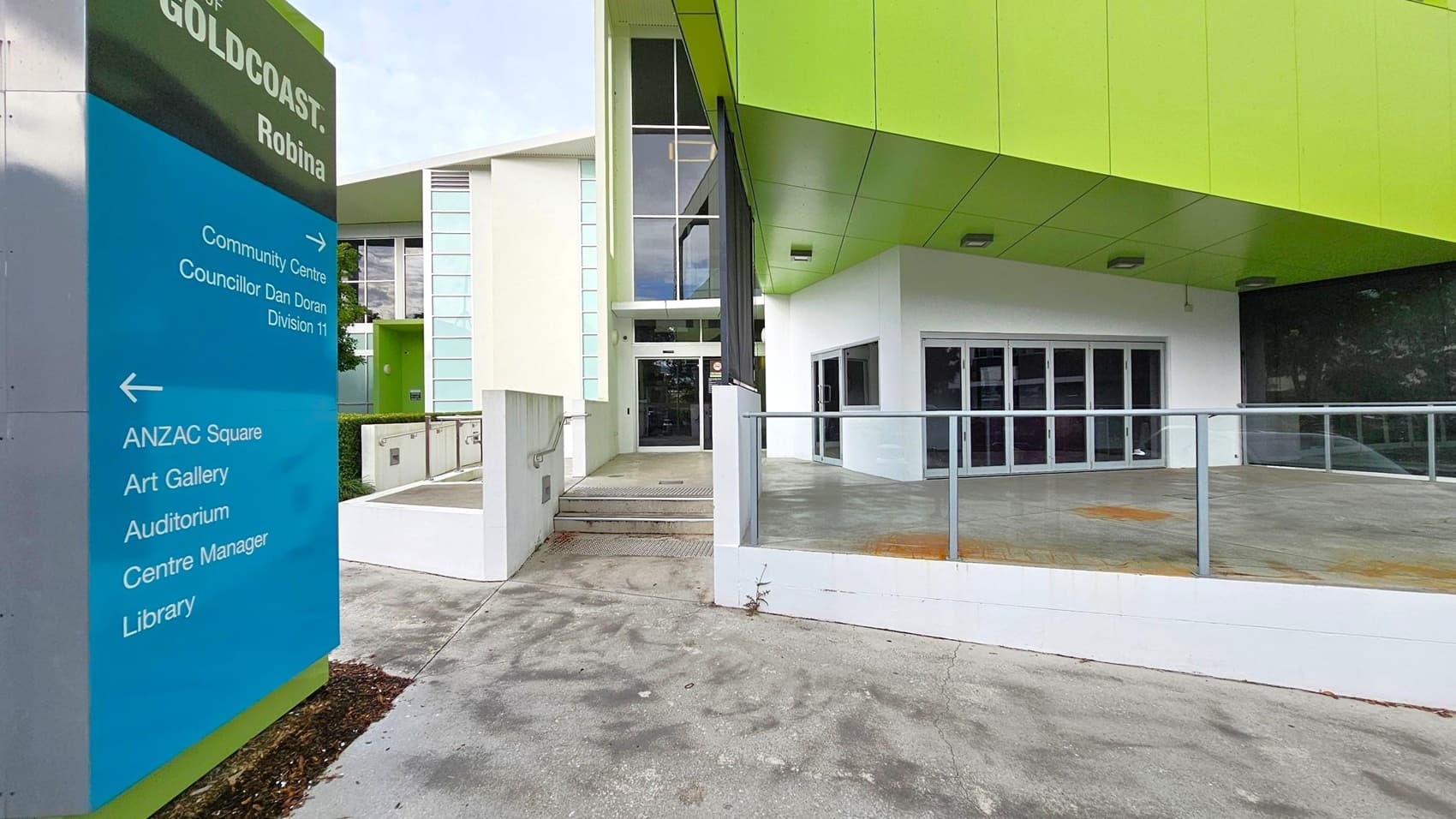 On the left, a blue and green sign reads "City of Gold Coast, Robina" with directions to various facilities like the community centre, ANZAC Square, art gallery, auditorium, centre manager, and library. In the centre, a modern building entrance features large glass doors and windows, with a green overhang above. The foreground has a concrete path leading to the entrance, bordered by a low white wall with a metal railing.
