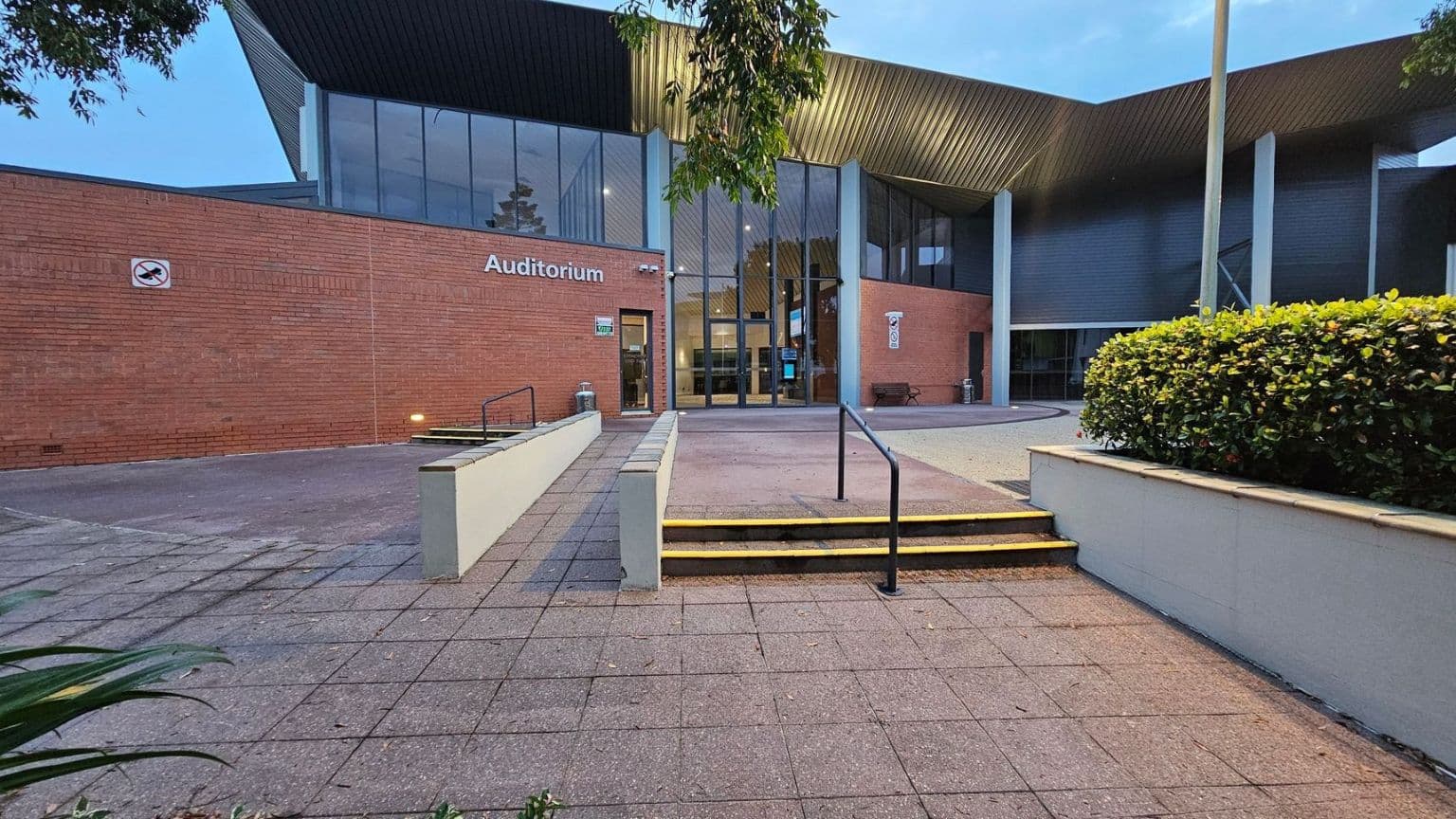 A modern building with a brick facade and large glass windows. The word "Auditorium" is visible on the left side. A no-smoking sign is mounted on the brick wall. The entrance features a set of steps with a handrail leading up to it. On the right, there is a neatly trimmed hedge. The foreground has a paved area with some fallen leaves. A tree branch is visible at the top left corner.