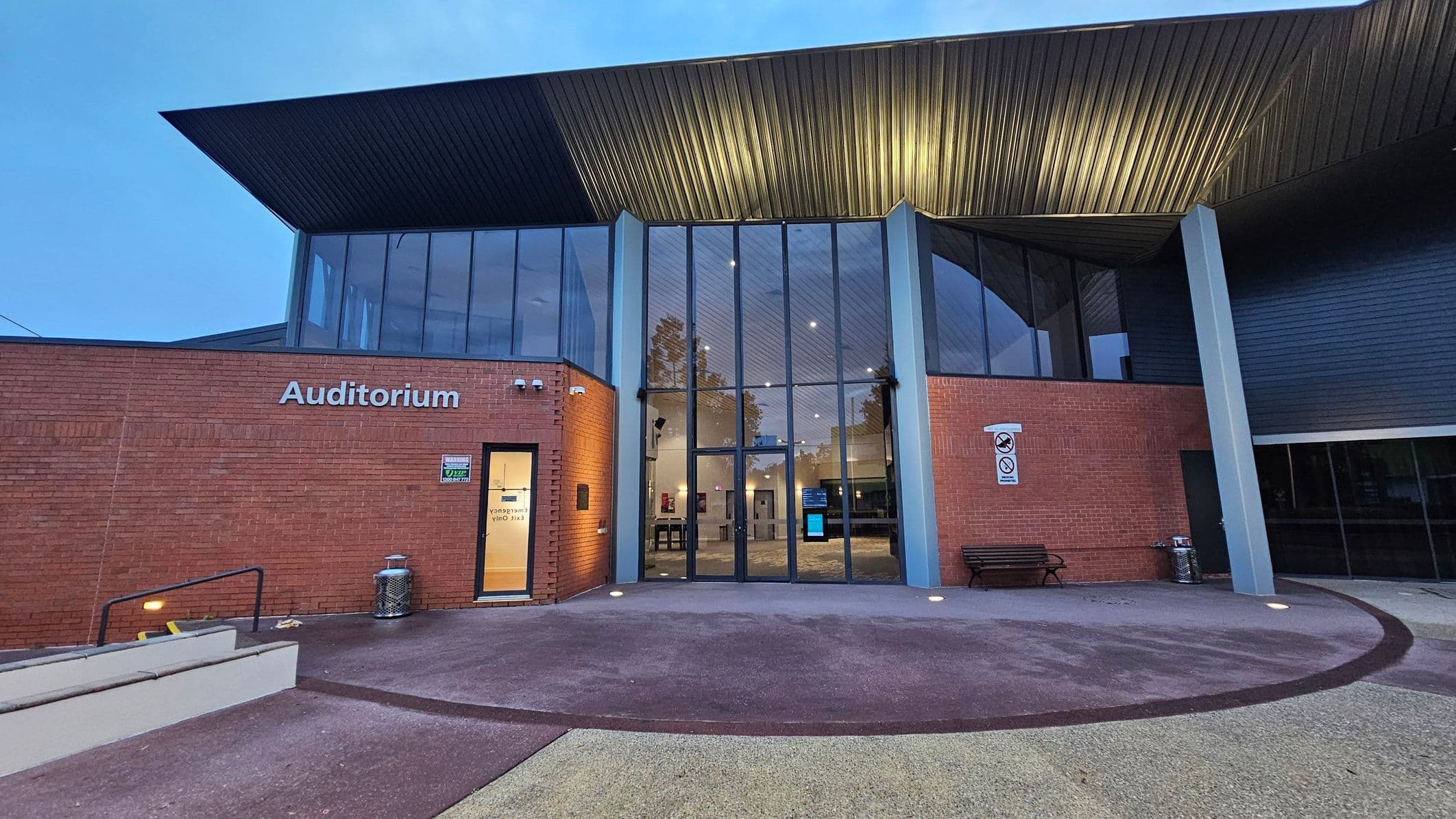A modern building with a large glass entrance in the centre, flanked by red brick walls. The word "Auditorium" is displayed on the left wall. A small door with a lit window is on the left, and a bench is on the right. The roof is angular and metallic, extending outward. The ground is a mix of smooth concrete and textured surfaces, with a ramp and railing on the left. A no-smoking sign is visible on the right wall.