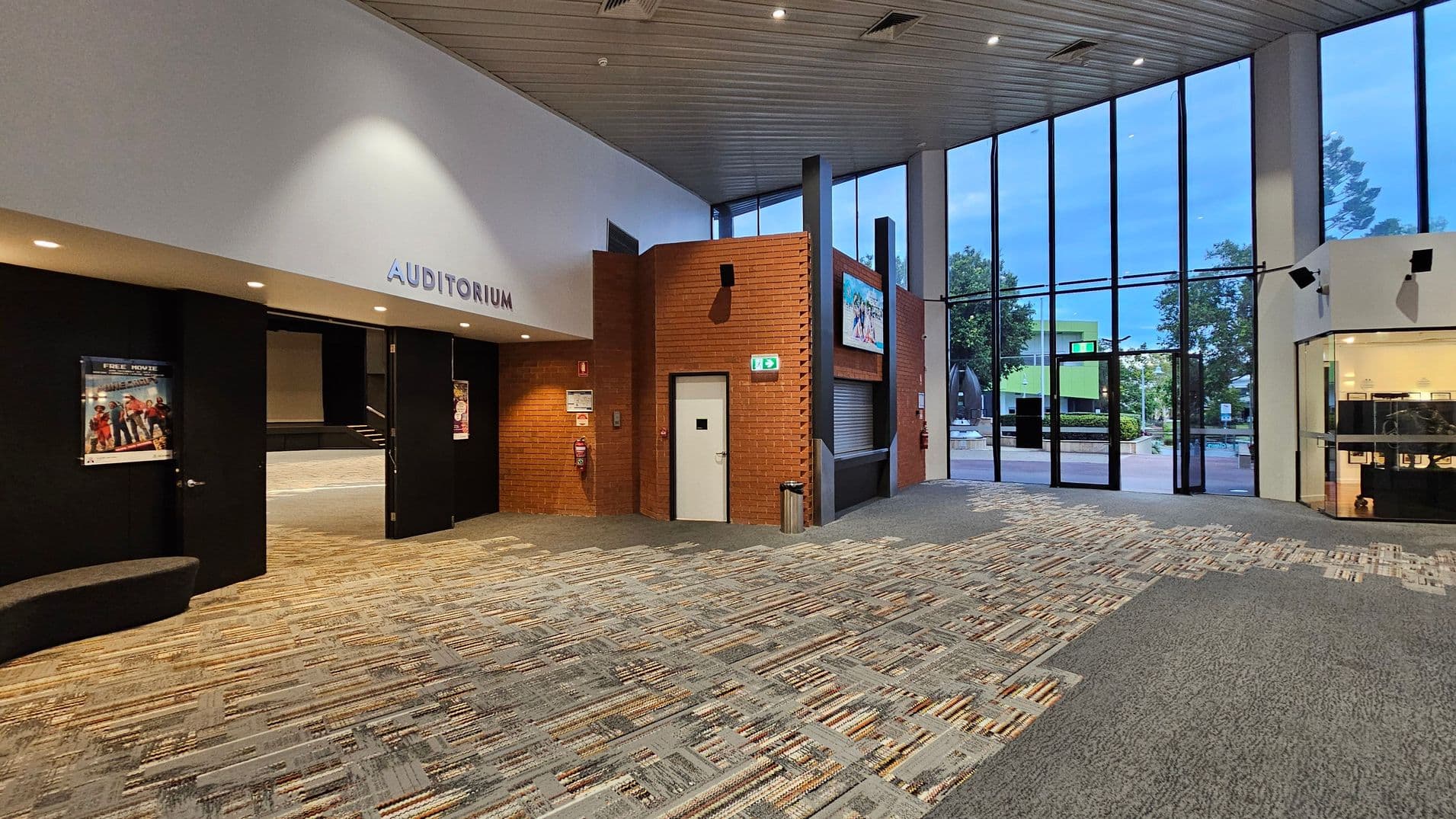 A spacious interior with a patterned carpeted floor. On the left, there is an entrance to an auditorium with black doors and a poster displayed. The centre features a brick wall with a white door and a digital screen above. On the right, large floor-to-ceiling windows reveal an outdoor view with trees and a building. The ceiling is lined with panels and spotlights.