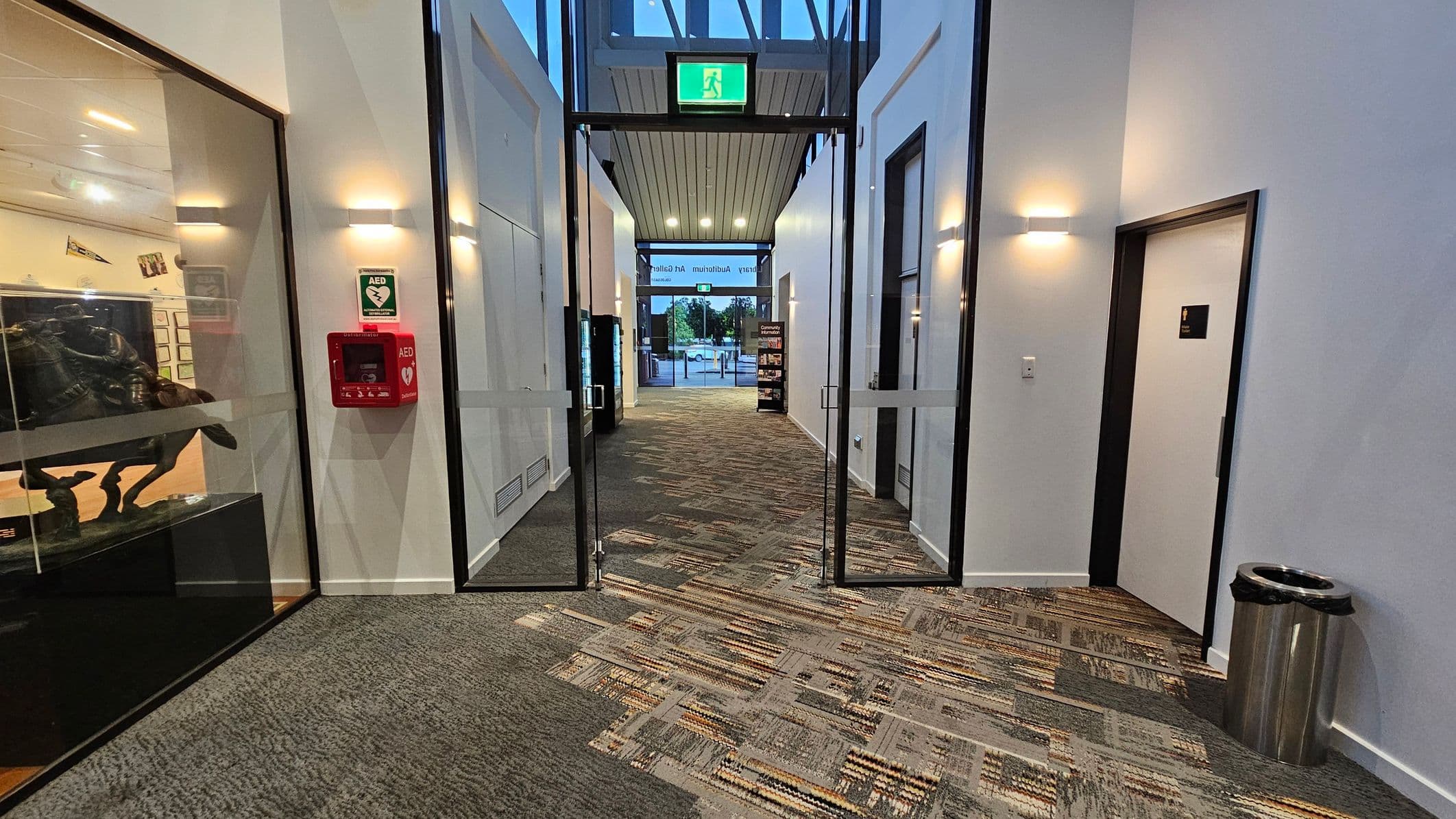 A hallway with a patterned carpet floor and white walls. On the left, there's a glass display case with a sculpture and a red AED box mounted on the wall. On the right, a door with a sign and a metal rubbish bin. The ceiling has recessed lighting and an exit sign above glass doors leading to an outdoor area. In the background, there's a view of trees and a building through the glass doors.
