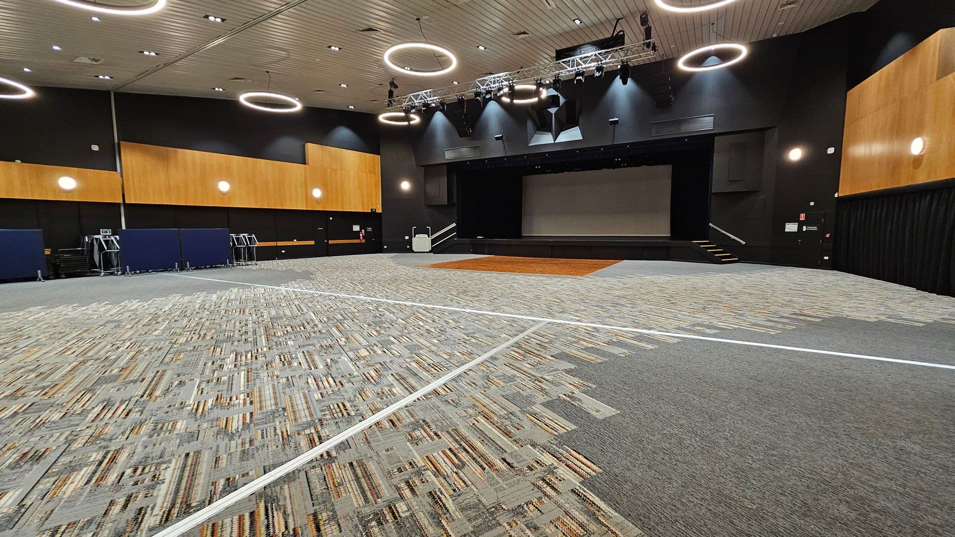 A large auditorium with a stage at the centre back. The floor is covered with a patterned carpet in shades of grey and green, with a wooden section near the stage. The walls are dark with wooden panels and circular lights. The ceiling is light-coloured with circular light fixtures. On the left, there are stacked chairs and equipment. The right side has a black curtain and an exit door.