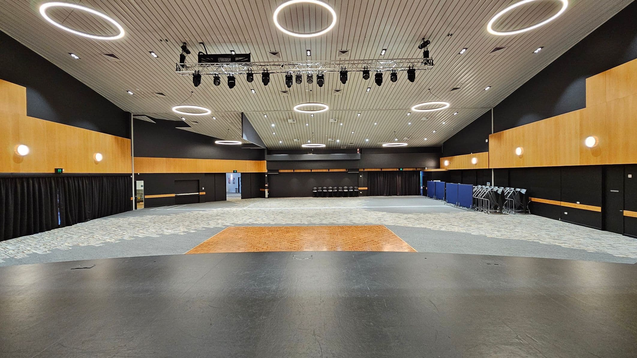 A large, empty hall with a high ceiling featuring circular light fixtures. The walls are a combination of black and wood panelling, with round wall lights. The floor is a mix of grey and patterned carpet, with a wooden section in the centre. On the right, there are stacked chairs and blue partitions. The ceiling has a grid of spotlights and a rig with stage lights. The left side has black curtains along the wall.