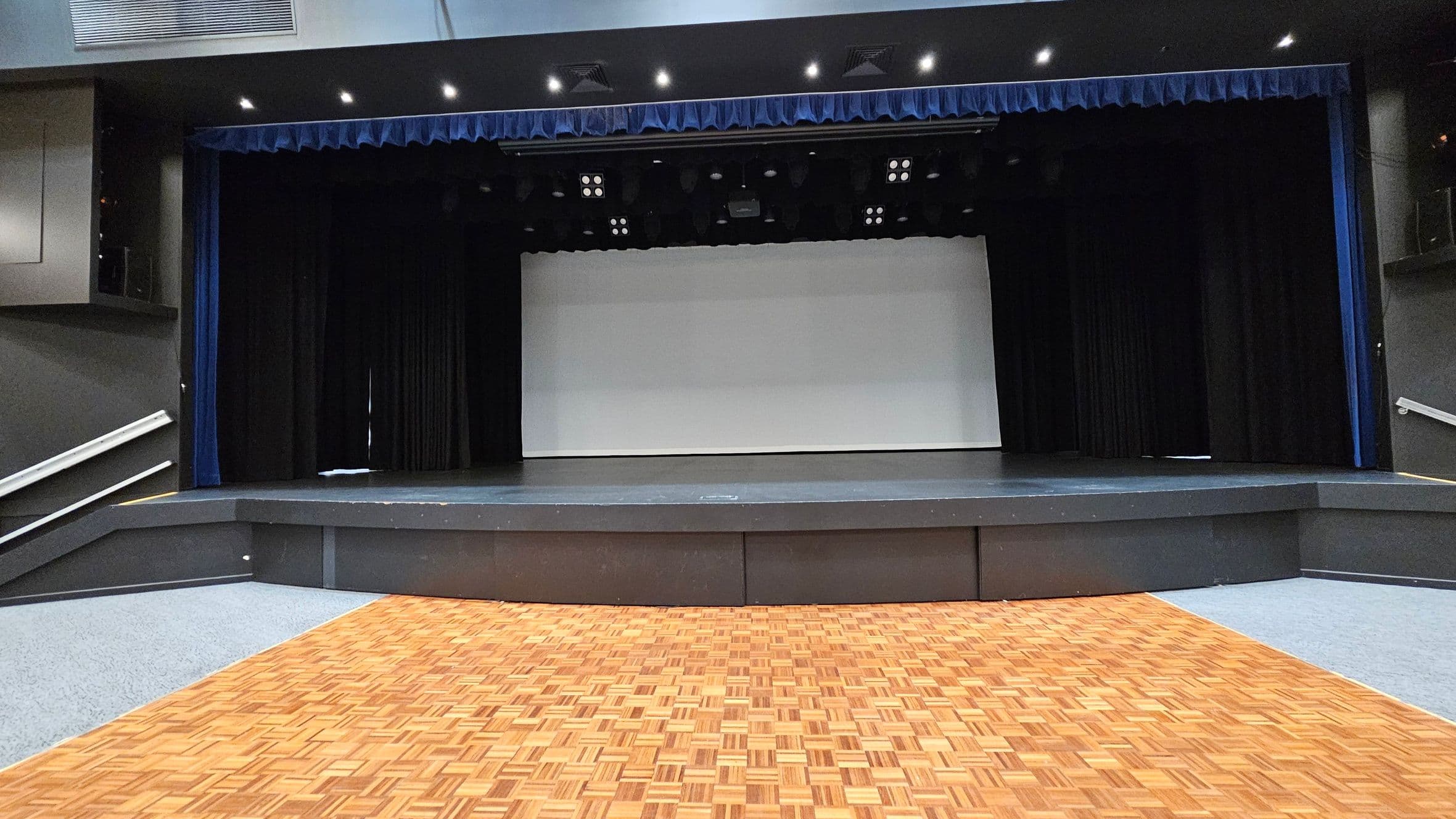 A stage with a wooden parquet floor in the foreground. The stage is elevated with a dark grey front and a large white screen in the centre. Black curtains frame the sides, and a blue valance runs along the top. The ceiling is equipped with multiple spotlights and ventilation panels. The left and right sides have angled ramps with handrails leading up to the stage. The surrounding walls are dark, complementing the stage setup.