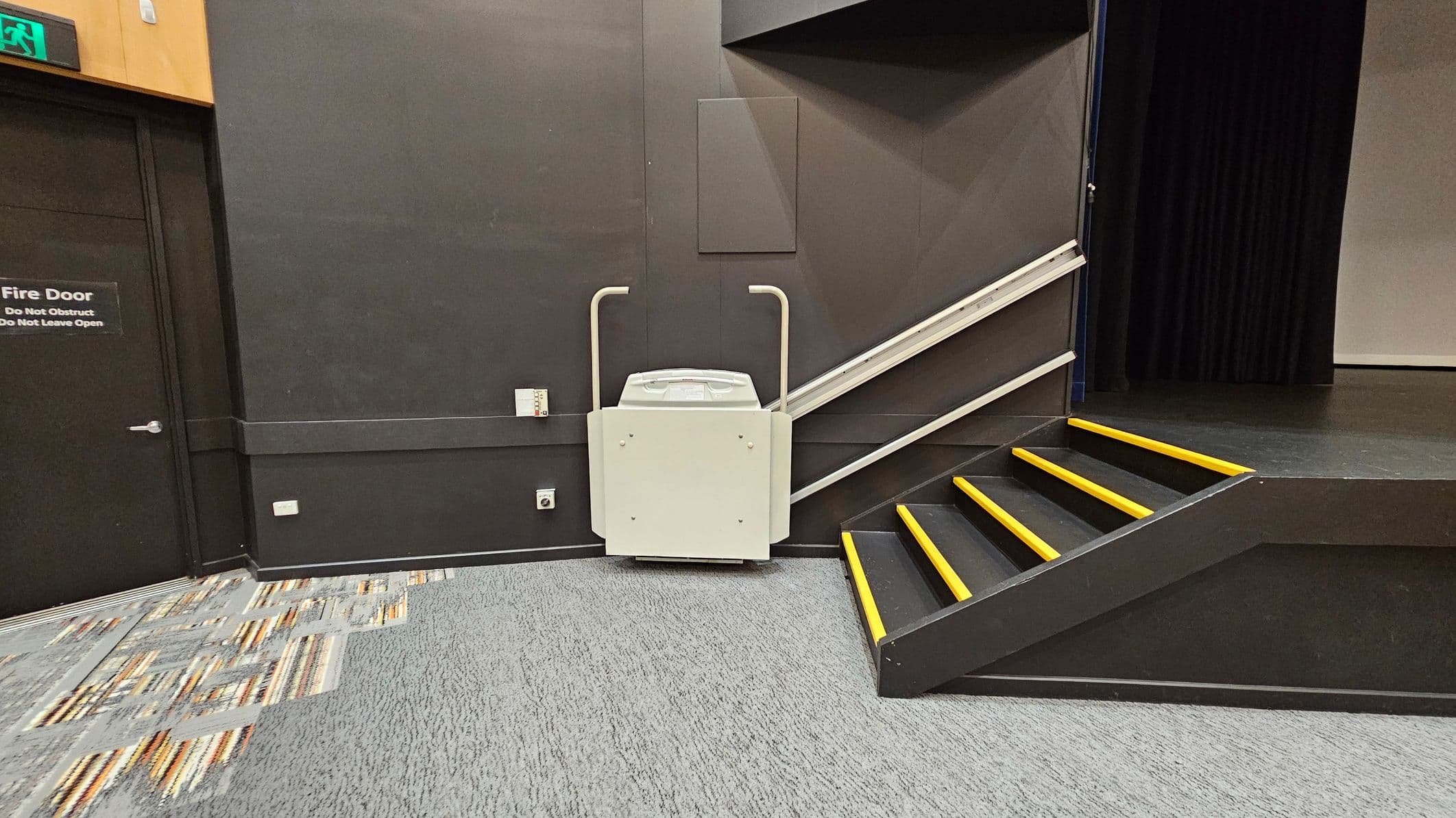 A room with a dark wall and a fire door on the left, marked with a sign. In the centre, a stair lift is attached to a sloped railing beside a set of black stairs with yellow-edged steps leading to a stage area on the right. The floor is covered with a mix of grey and patterned carpet tiles.
