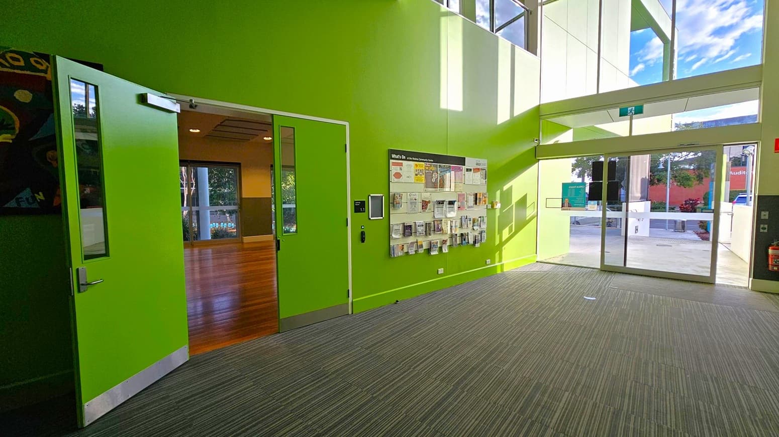 A bright green interior space with a carpeted floor. On the left, a green door is open, revealing a room with wooden flooring. In the centre, a wall-mounted display holds various brochures and pamphlets. On the right, large glass doors lead outside, allowing natural light to flood in. The ceiling is high with large windows above, enhancing the airy feel of the space.