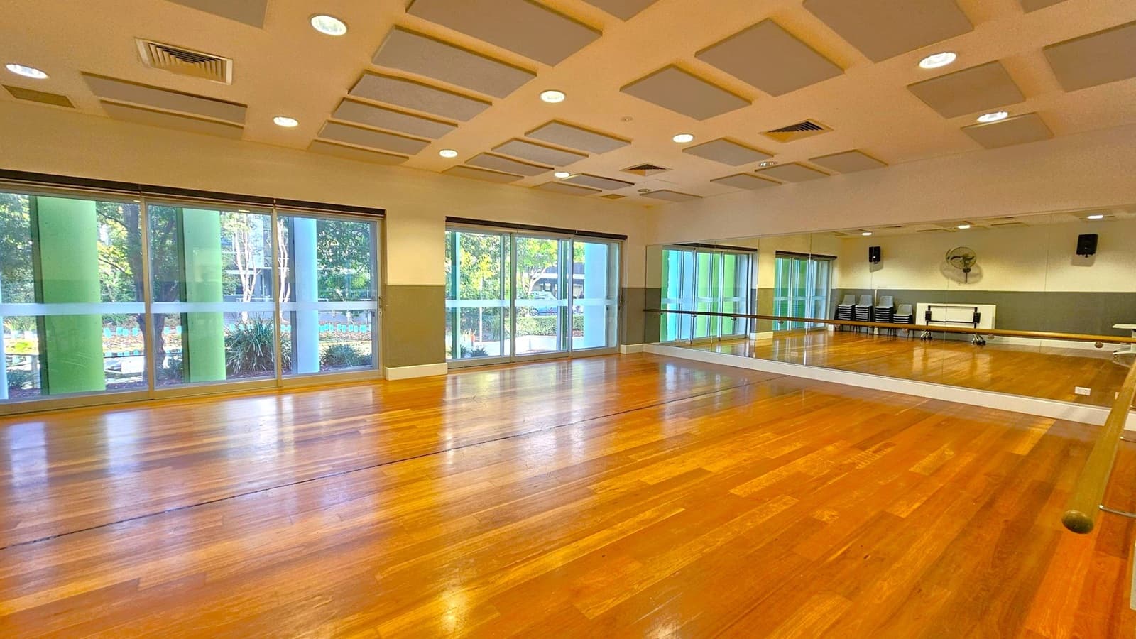 A spacious dance studio with a polished wooden floor. On the right, a large mirror spans the wall, reflecting the room. The left side features large windows with green pillars outside, allowing natural light to enter. In the background, stacked chairs and a wall-mounted fan are visible. The ceiling has recessed lighting and acoustic panels.