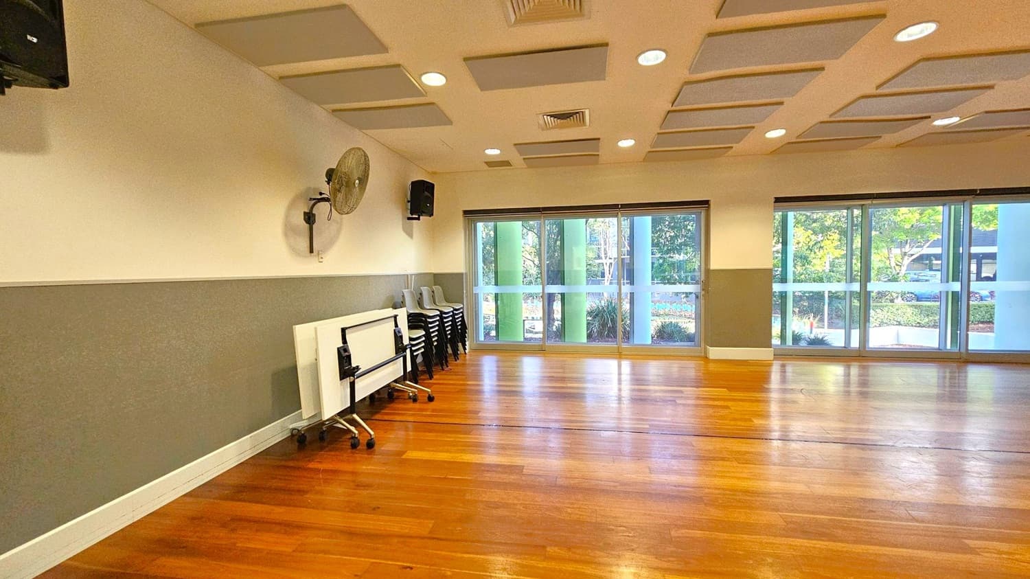 A spacious room with wooden flooring and a ceiling featuring recessed lighting and acoustic panels. On the left, a wall-mounted fan and a stack of chairs are visible, along with foldable tables on wheels. The right side has large windows with a view of greenery outside. The walls are painted in two tones, with the lower half in grey and the upper half in white.