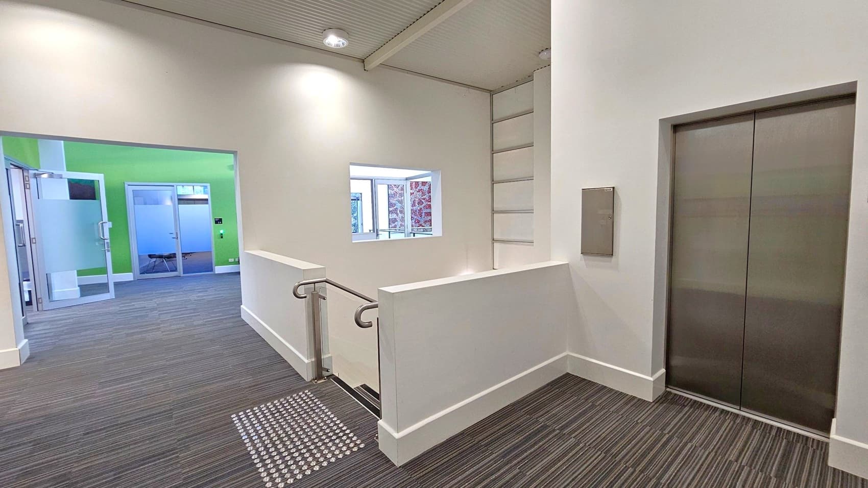 A modern indoor space with a grey carpeted floor. On the right, there is a closed lift with metallic doors. In the centre, a staircase with a white railing leads downwards. On the left, a corridor with a green wall and glass doors is visible. The ceiling is light-coloured with recessed lighting. A small window is set in the wall above the staircase, allowing natural light to enter.