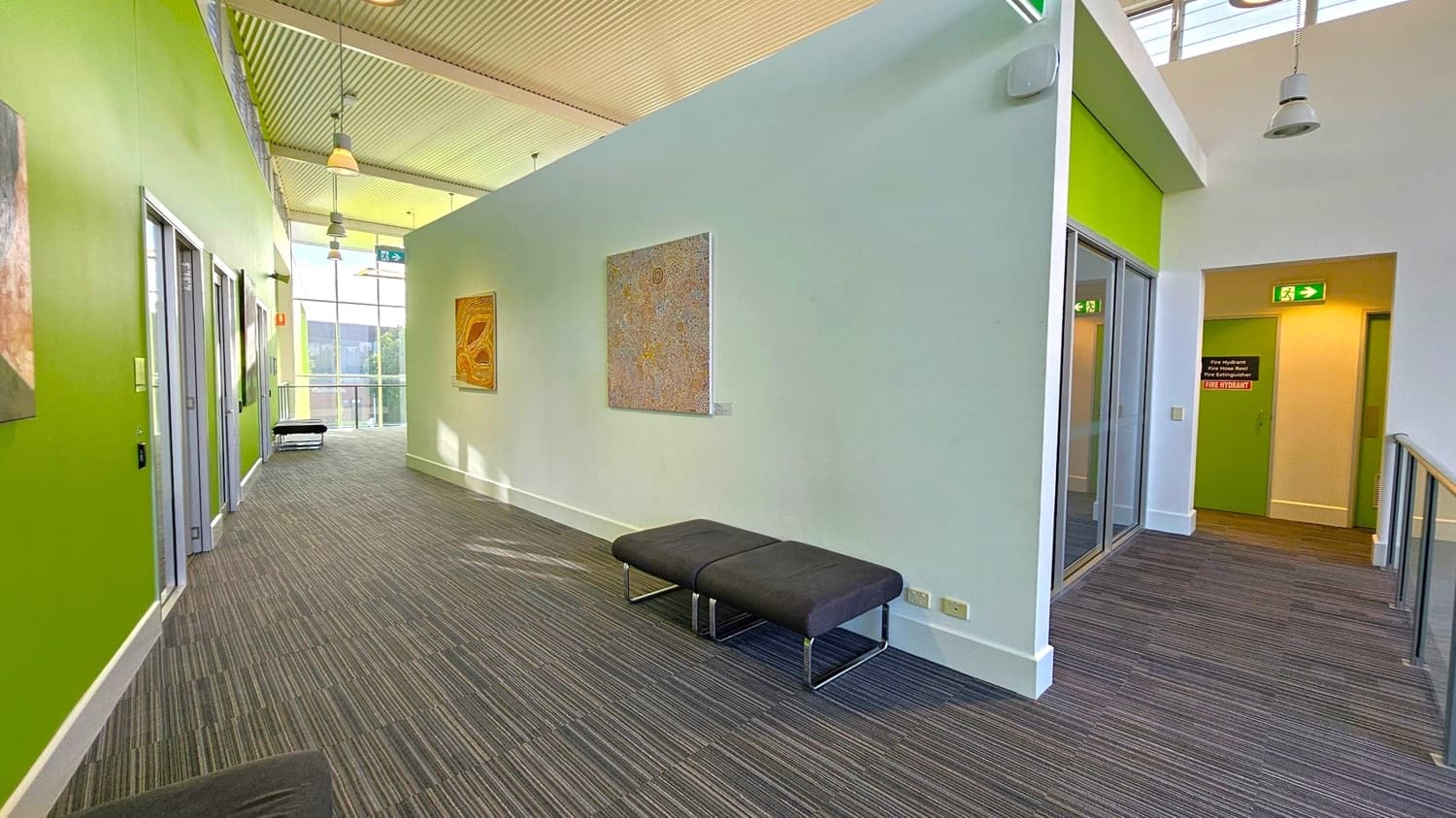 A corridor with green walls on the left and a light grey wall on the right. The floor is covered with striped carpet. On the left, there are several doors with glass panels and a bench at the end. The right wall features two pieces of artwork and another bench. The ceiling is wooden with hanging lights. An exit sign is visible above a door on the right. Large windows at the end of the corridor allow natural light to illuminate the space.