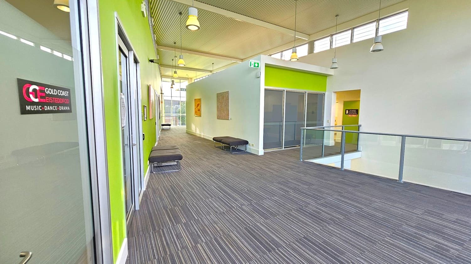 A modern hallway with a grey carpeted floor and a high ceiling featuring hanging lights. On the left, there's a glass door with a sign for "Gold Coast Eisteddfod Music Dance Drama" on a lime green wall. The right side has a glass railing overlooking a lower level. In the middle, there are black benches along the walls, which are adorned with framed artworks. The far end of the hallway is brightly lit by large windows.