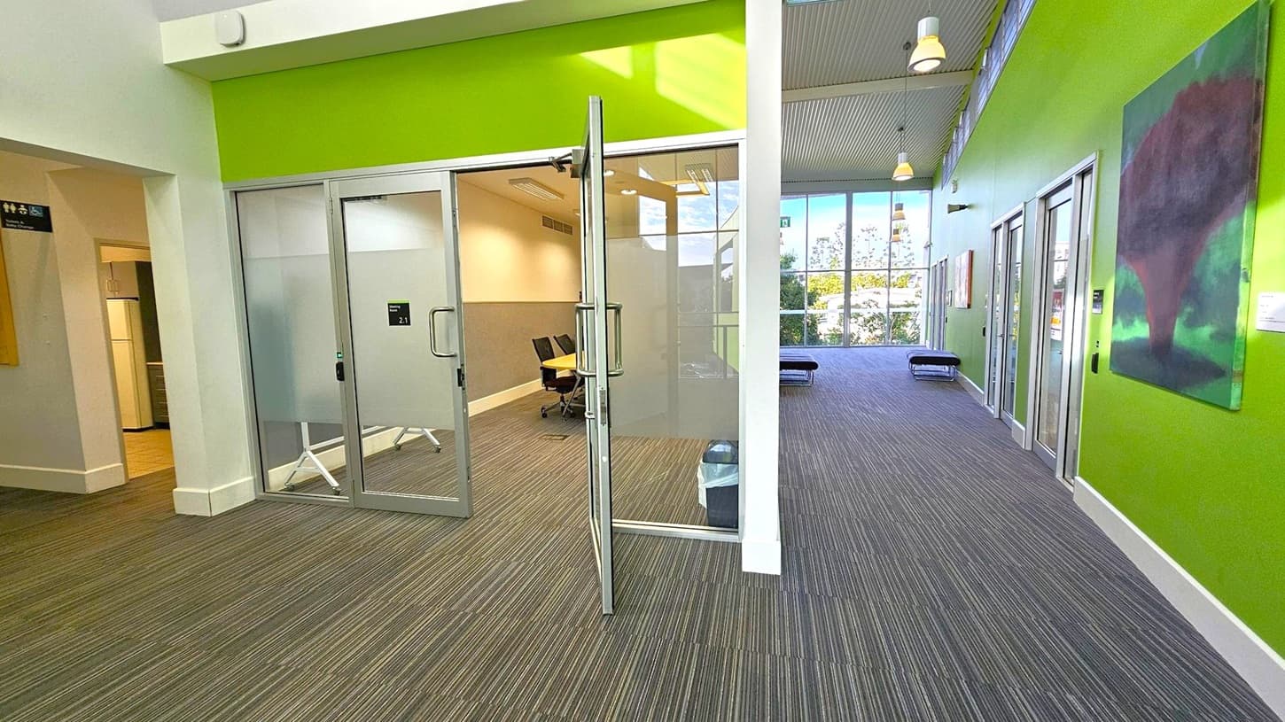 A modern interior space with a green accent wall on the right. Glass doors lead to a room with chairs and a table. The floor is covered with striped carpet. On the right wall, there's a large abstract painting. The ceiling has hanging lights, and large windows in the background allow natural light to enter. On the left, there's an open doorway leading to another area.