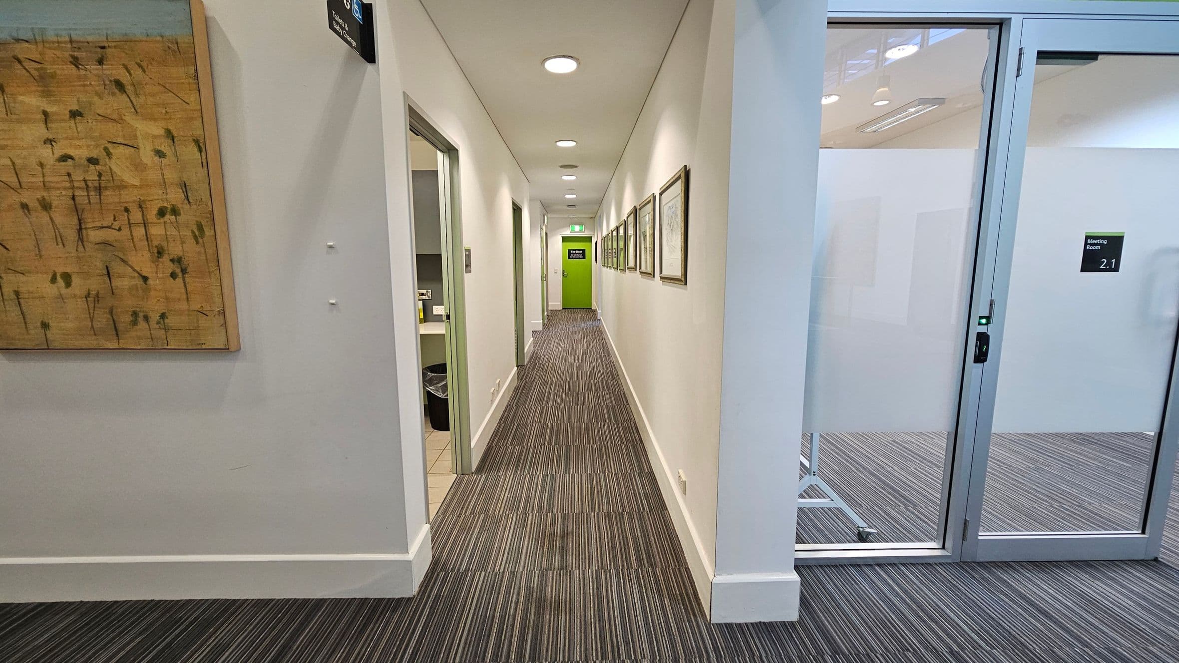 A corridor with a striped carpet floor and white walls. On the left, there's a wooden artwork and an open door leading to a room with a bin visible. On the right, there's a glass door labelled "Meeting Room 2.1". The corridor is lined with framed pictures on the right wall, leading to a bright green door at the end. The ceiling has recessed lighting.