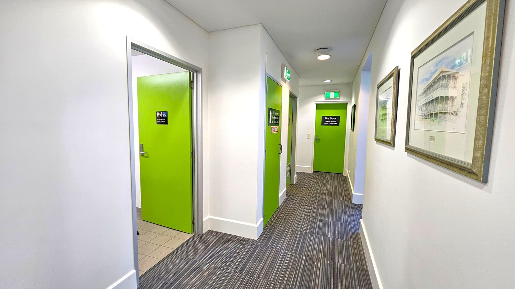 A hallway with a striped carpeted floor and white walls. On the left, there is an open door with a bright green colour, leading to a tiled room. Further down the hallway, two more closed green doors are visible, each with signs. On the right wall, there are framed pictures hanging. The ceiling is white with recessed lighting and an emergency exit sign above the far door.
