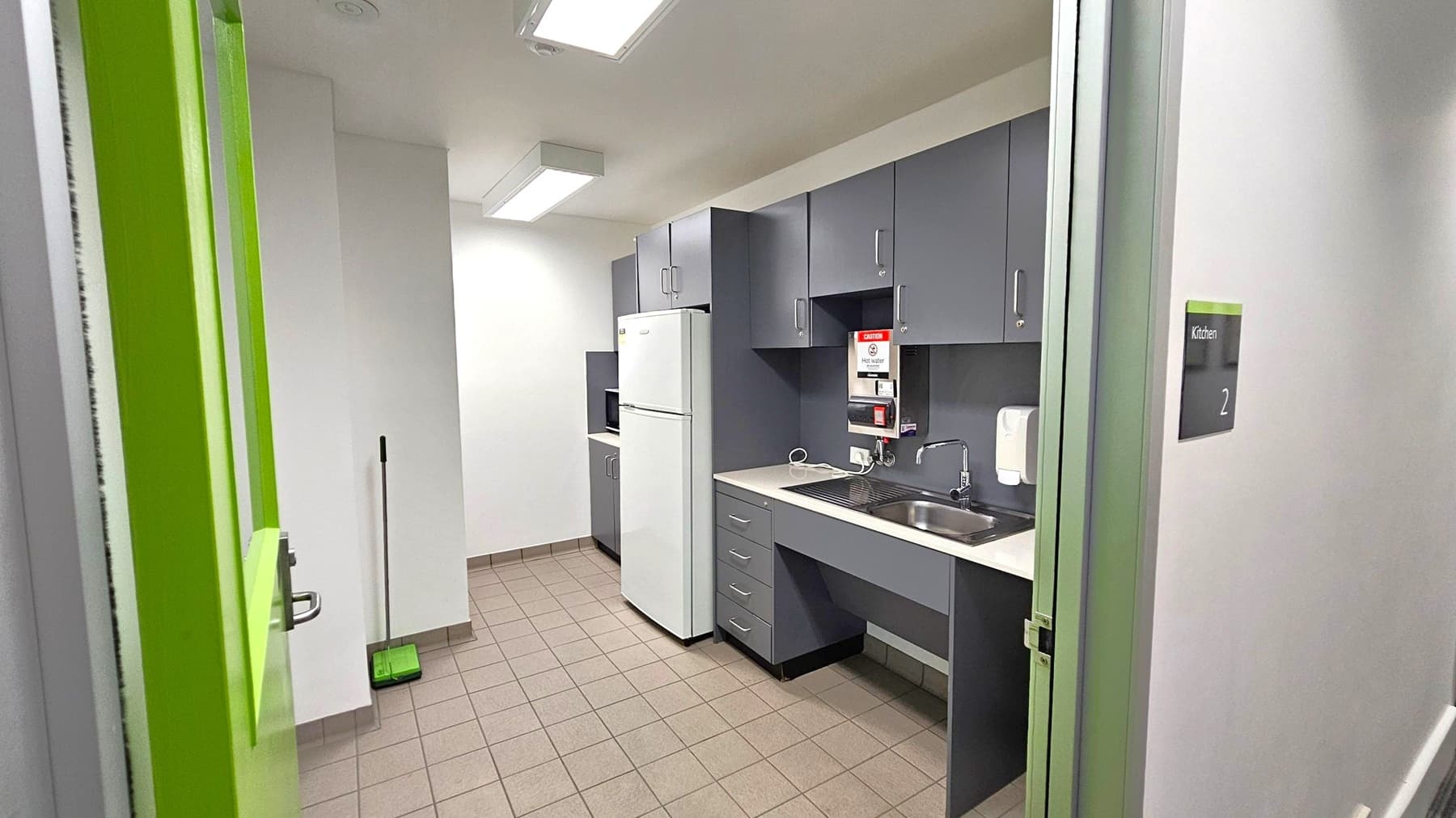 A small kitchen with grey cabinets and a white refrigerator on the right. A sink with a tap is set into the countertop, and a microwave is visible. A green door is partially open on the left, with a broom and dustpan leaning against the wall. The floor is tiled in a light brown colour, and the ceiling has fluorescent lights. A sign on the wall reads "Kitchen 2".