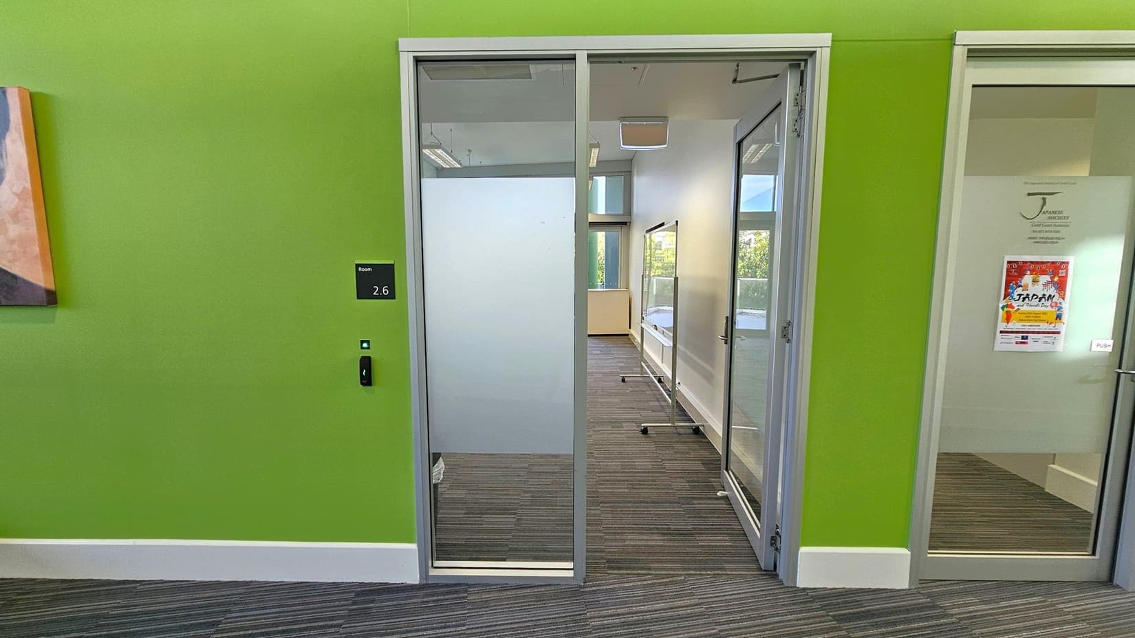 A room with a green wall and a glass door labelled "2.6" on the left. The door has a frosted lower section and a clear upper section. Inside, there's a carpeted floor with a striped pattern and a window at the far end. To the right, another glass door with a poster on it. A portion of a painting is visible on the far left.