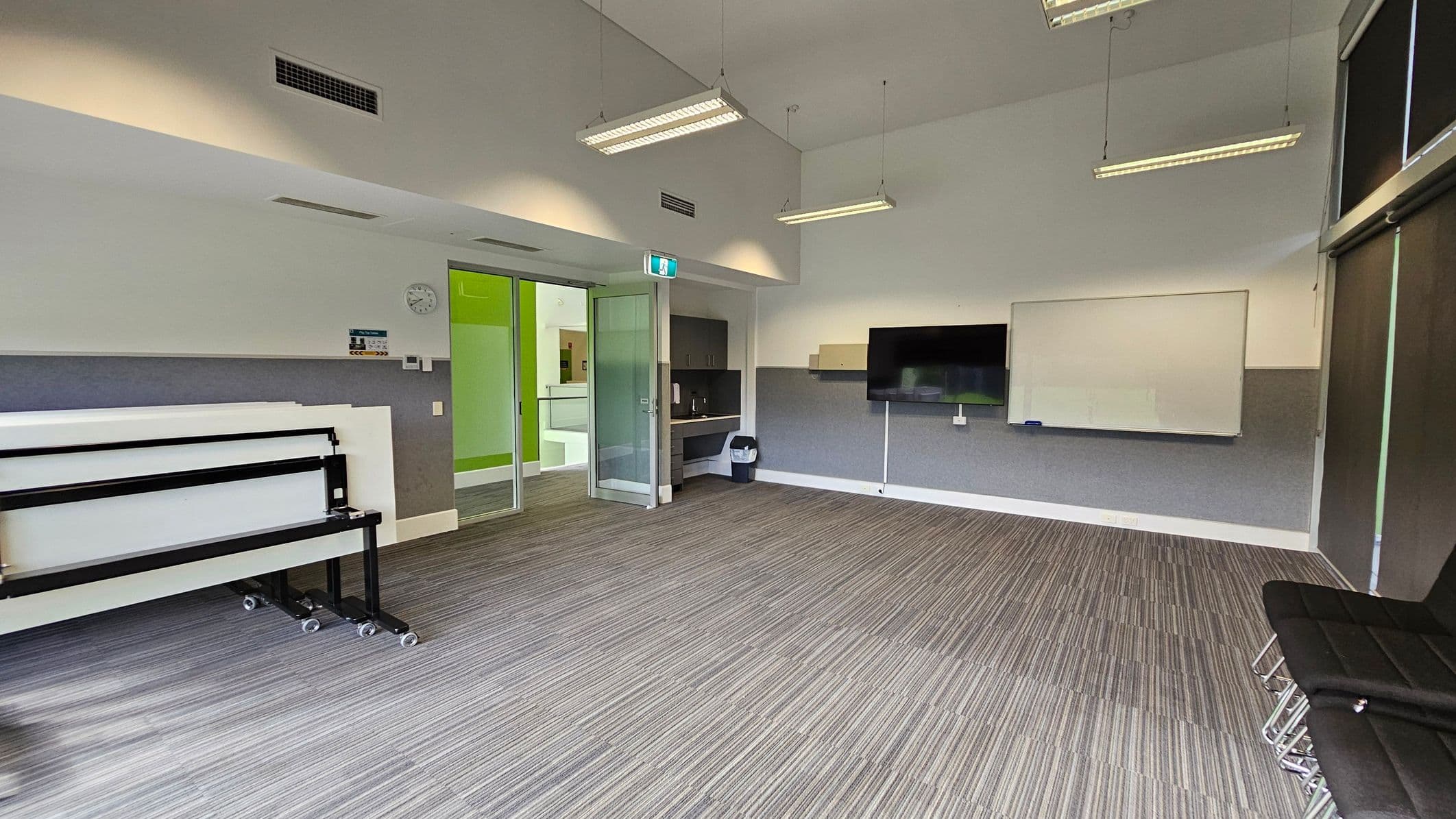 A modern classroom with a striped carpeted floor and a high ceiling with hanging lights. On the left, there are stacked tables on wheels. In the centre, a glass door leads to a hallway with green walls. A wall-mounted TV and a whiteboard are on the right wall. Below the TV, there is a small counter with a bin underneath. A stack of black chairs is in the right foreground. A clock and some controls are mounted on the left wall.