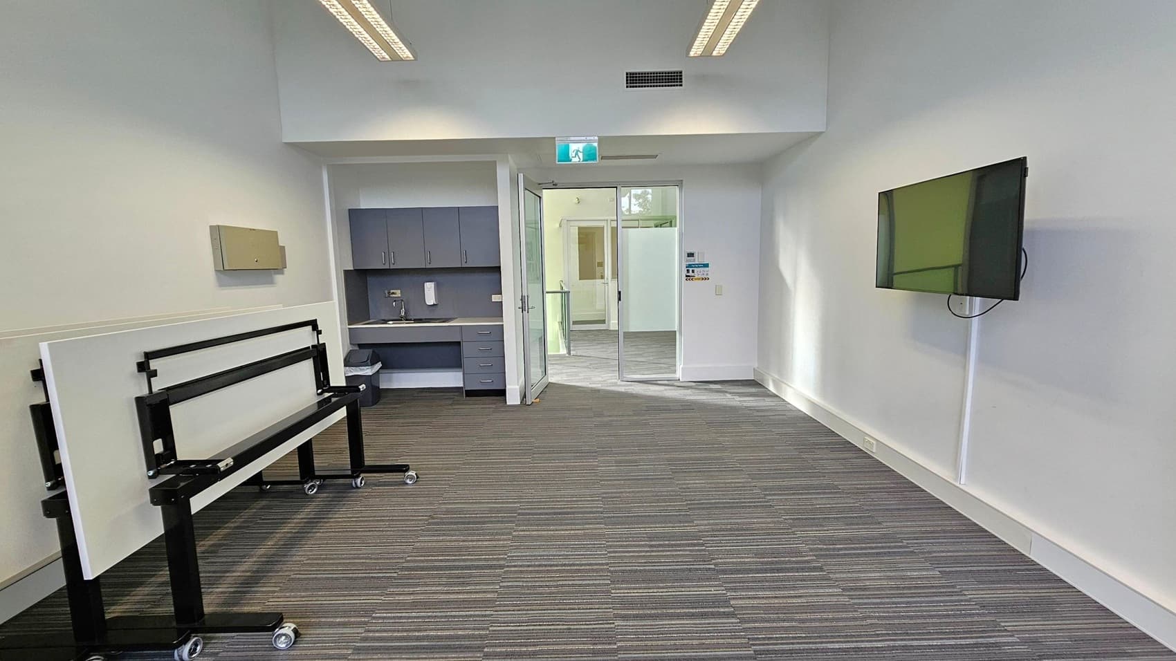A room with a striped carpeted floor and a white ceiling with recessed lighting. On the left, there are stacked tables on wheels and a wall-mounted cabinet. In the centre, a doorway with a glass door leads to another area, with an exit sign above. On the right, a flat-screen TV is mounted on the wall. A small kitchenette with cabinets, a sink, and a soap dispenser is visible near the doorway.