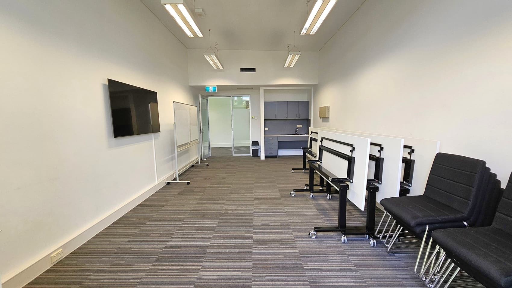 A narrow room with a striped carpeted floor and a white ceiling with fluorescent lights. On the left wall, a large flat-screen TV is mounted, and a whiteboard is positioned nearby. On the right, several black chairs are stacked, and foldable tables are lined up. At the far end, there is a kitchenette with grey cabinets and a sink, and a glass door with an exit sign above it.