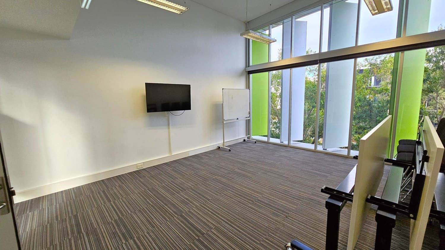 A modern room with a striped carpeted floor. On the left wall, a flat-screen TV is mounted, and a whiteboard on wheels is nearby. The right side features large floor-to-ceiling windows with vertical green and white panels, offering a view of trees outside. In the foreground, there are stacked tables with black legs. The ceiling has rectangular fluorescent lights.