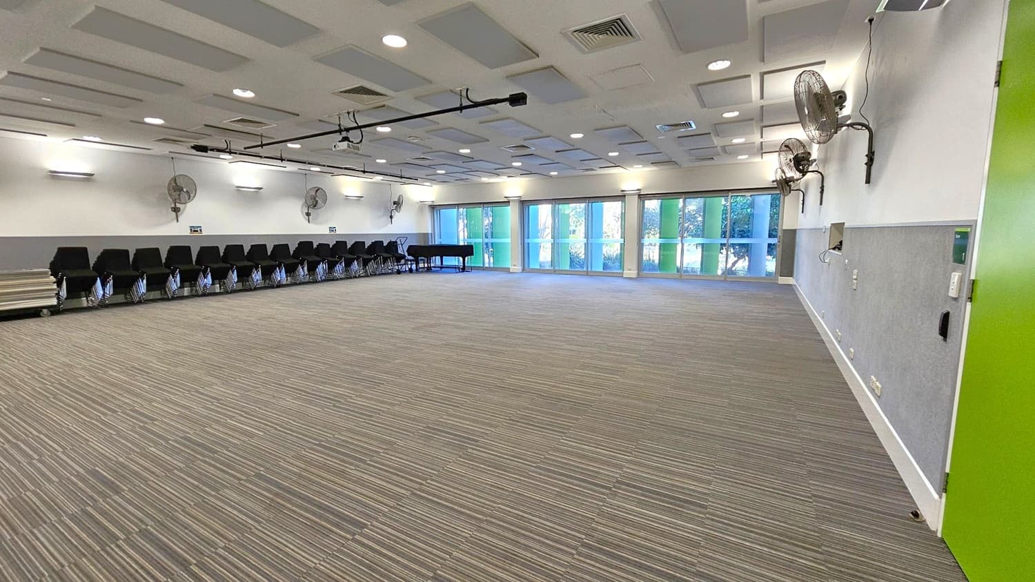 A spacious room with a striped carpeted floor. On the left, there are rows of black chairs lined up against the wall, with several wall-mounted fans above them. A small stage is visible at the far end. On the right, a green door is partially open. The back wall features large windows with blue and green panels, allowing natural light to enter. The ceiling has a grid pattern with recessed lighting and ventilation panels. A black piano is near the windows.