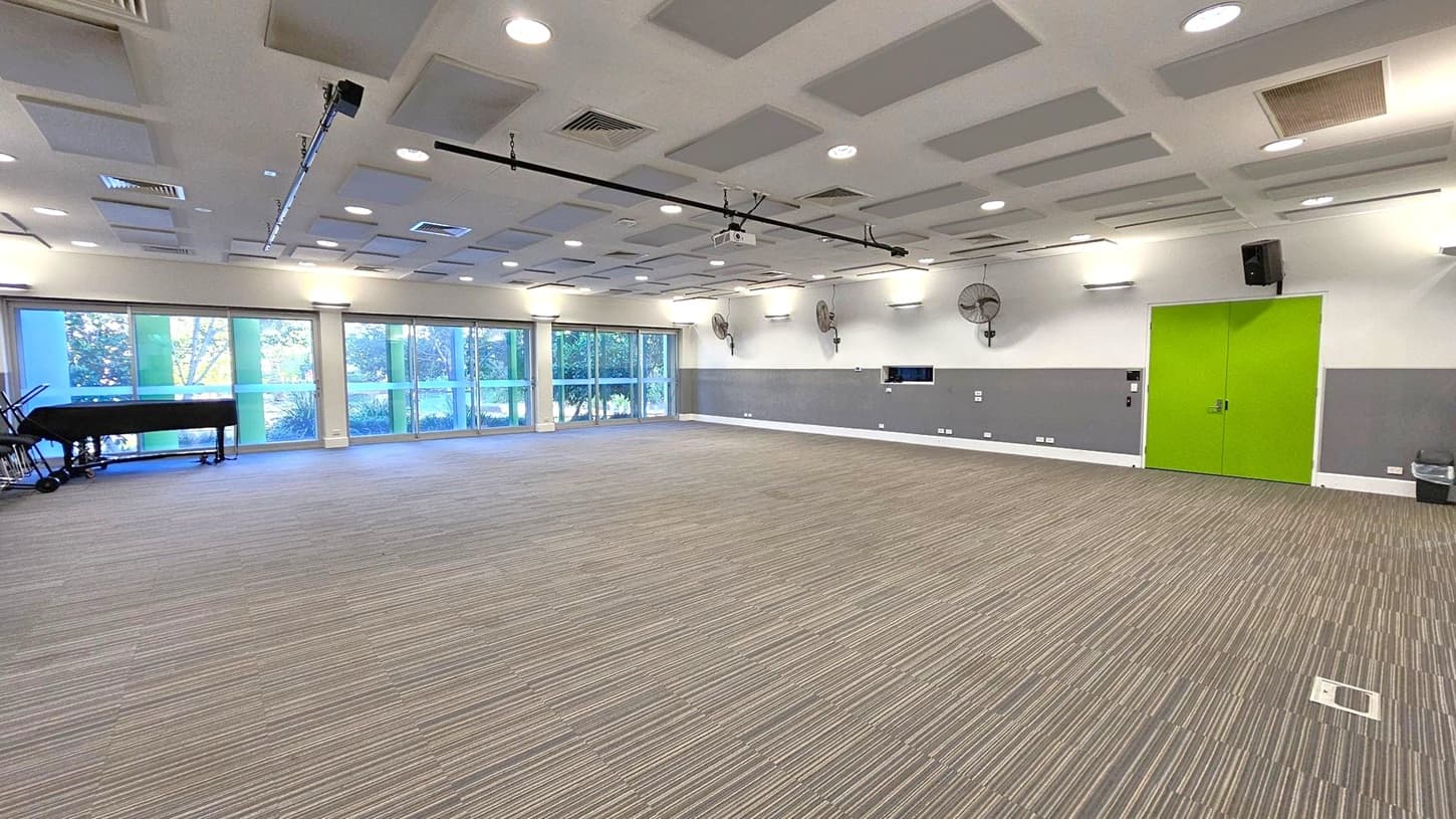 A spacious room with a striped carpeted floor. On the left, large windows with green-tinted glass panels allow natural light to enter, and a black grand piano is positioned near the windows. The right side features a bright green double door. The walls are grey and white, with mounted fans and speakers. The ceiling is fitted with square acoustic panels and recessed lighting.