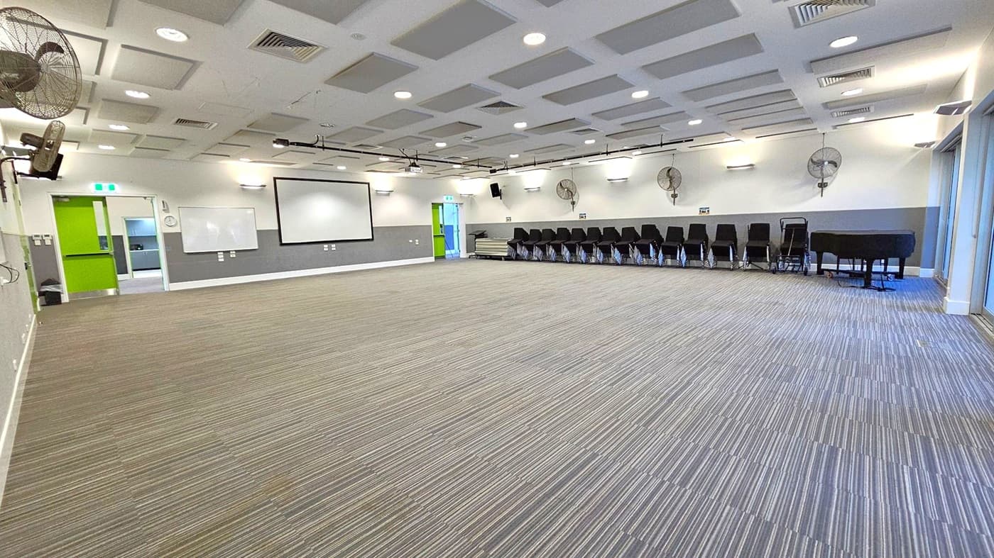 A spacious room with a striped carpeted floor. On the left, there are green double doors and a whiteboard mounted on the wall. In the centre, a large projection screen is visible. On the right, a row of stacked black chairs lines the wall, next to a black piano. The ceiling features recessed lighting and ventilation panels, with wall-mounted fans along the right side. The room is well-lit with a modern design.