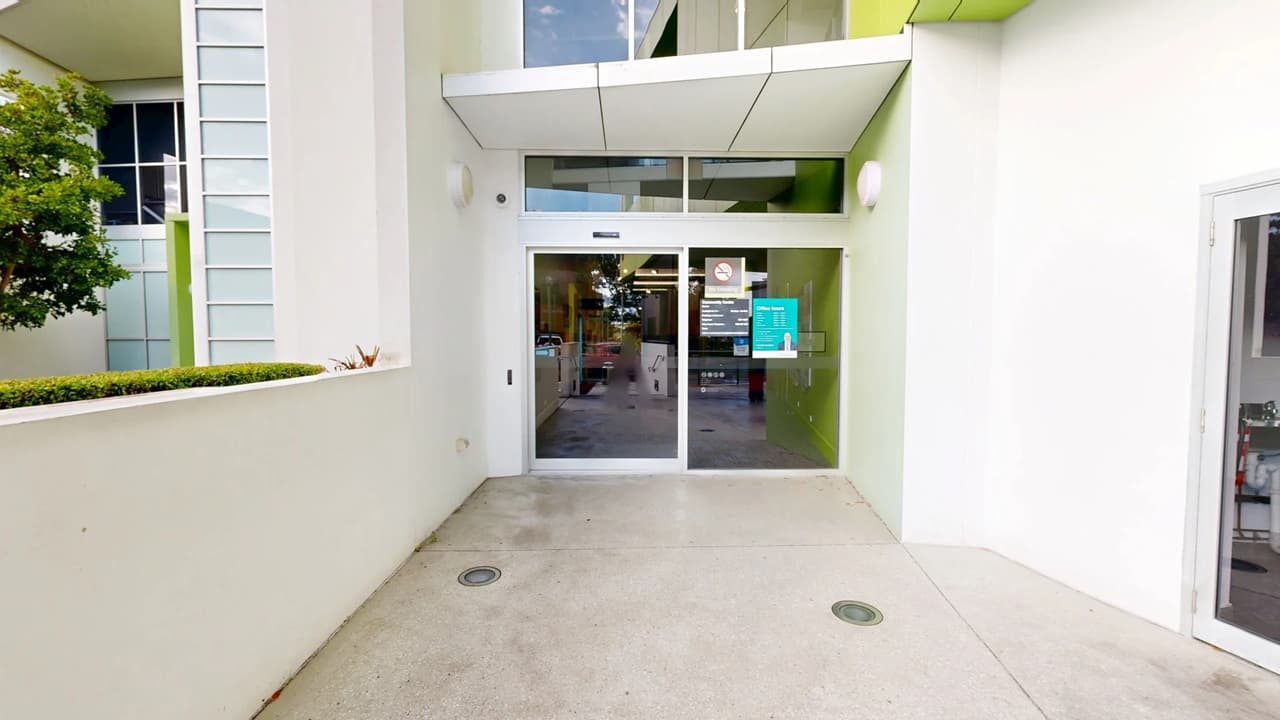A modern entrance with glass sliding doors in the centre, featuring a sign and a no-smoking symbol. The building exterior is white with green accents. On the left, there's a small tree and a row of windows. The floor is concrete with two circular drains. The ceiling is white with recessed lighting. On the right, part of another glass door is visible.