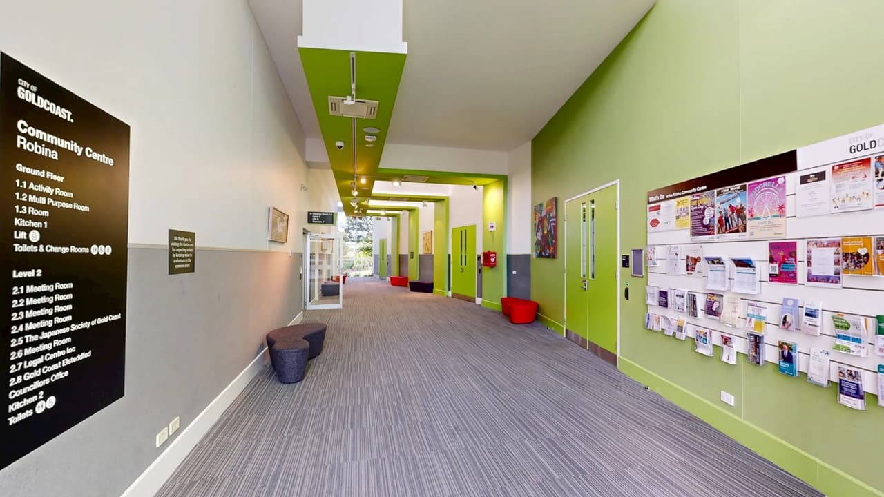 A hallway with a grey carpeted floor and a green ceiling. On the left, a black sign lists rooms in the Community Centre Robina. Further down, a glass door and a few chairs are visible. On the right, a green wall features a display of brochures and posters, with green doors and a red fire extinguisher. The corridor is well-lit with overhead lights, and there are red and black seating options along the walls.