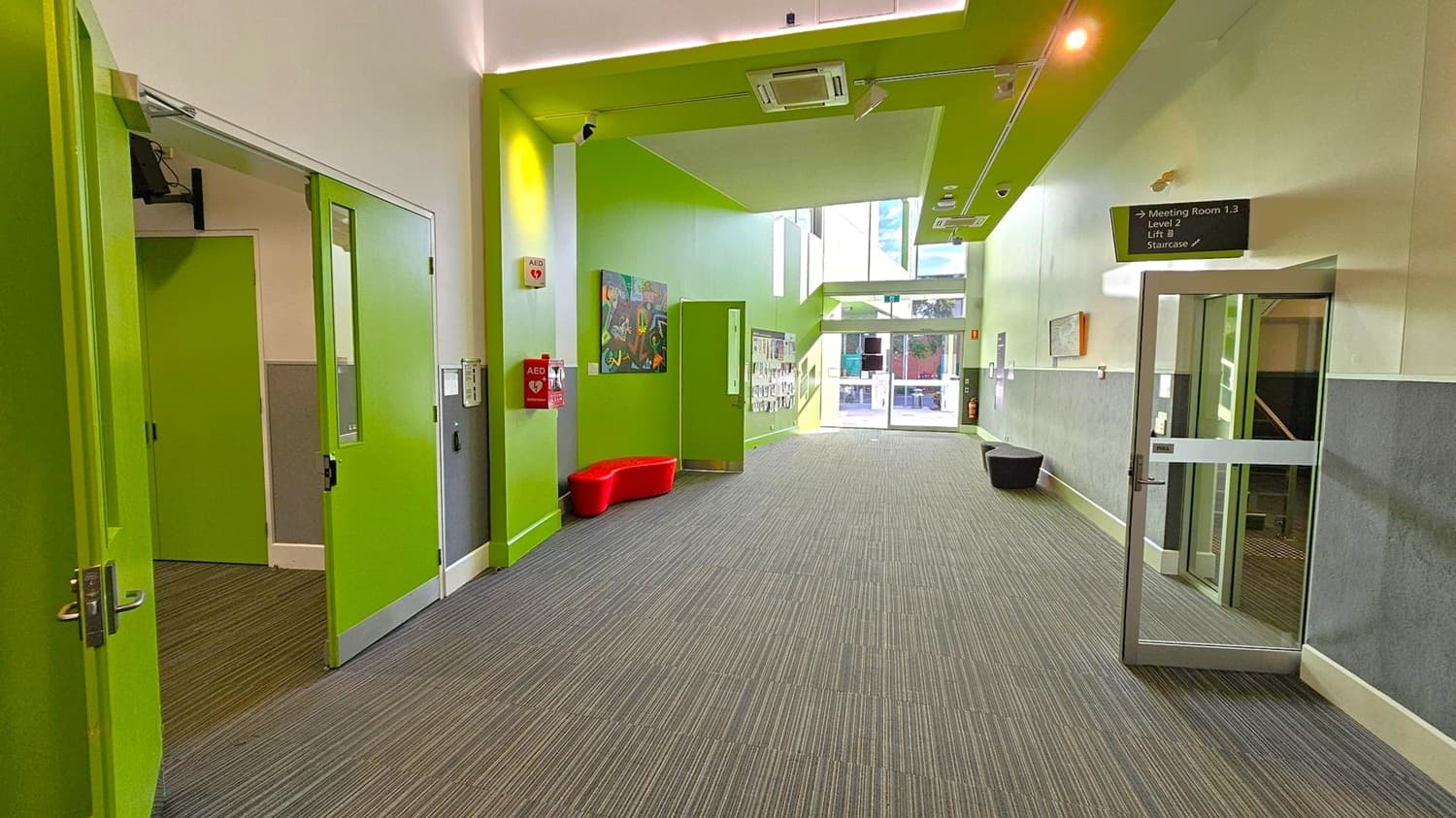A hallway with bright green walls and doors. On the left, there are open green doors leading to another room. In the centre, a red bench is placed against the wall, with a colourful painting above it. An AED device is mounted on the wall nearby. The floor is covered with grey carpet tiles. On the right, a glass door leads to a staircase. The ceiling has green panels with lights and an air conditioning unit. At the far end, large windows allow natural light to enter.