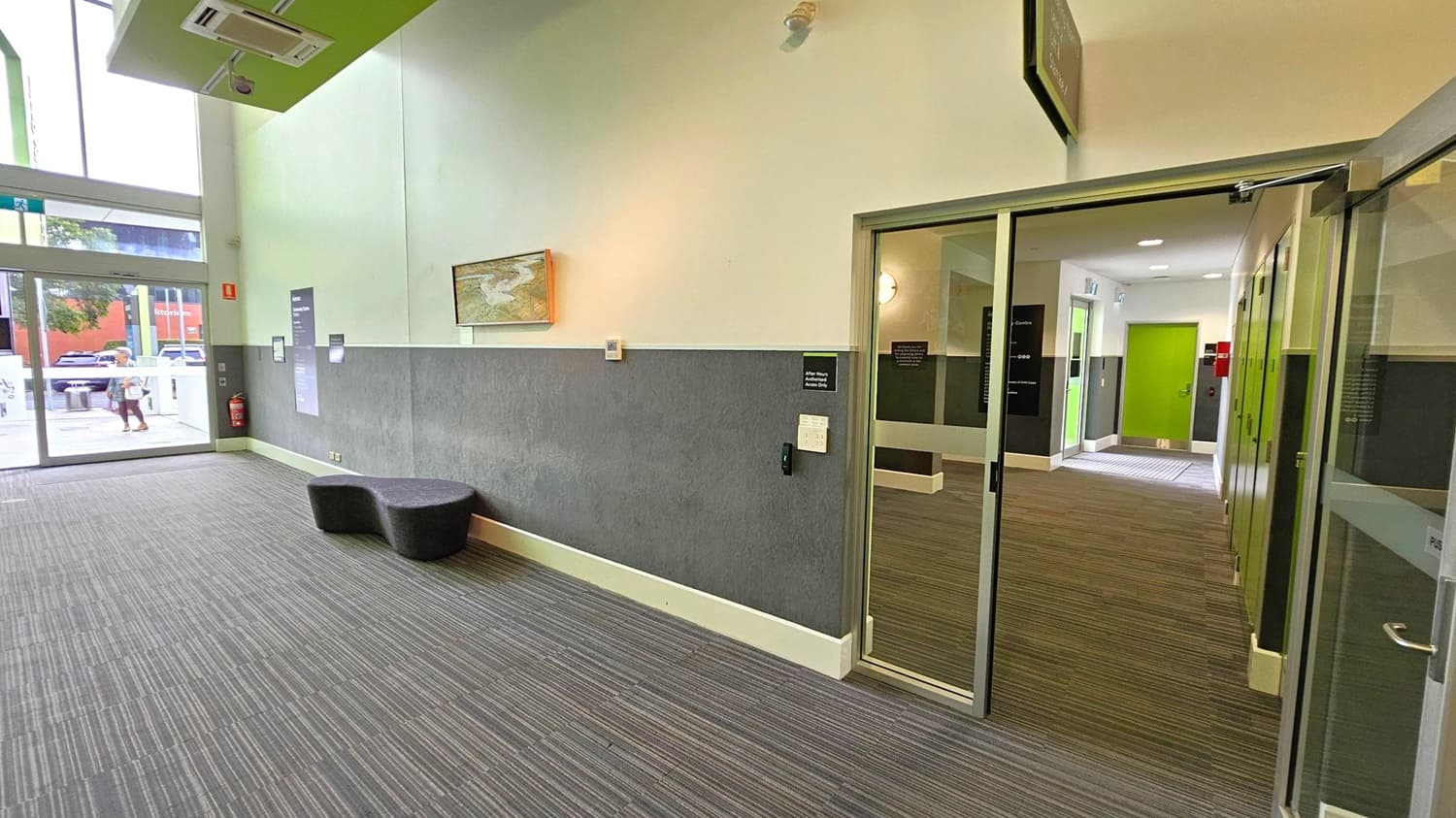 A modern interior space with a grey carpeted floor. On the left, large glass doors lead outside, where a person and parked cars are visible. A black, curved bench sits against the wall, which is half grey and half white. A painting hangs above the bench. On the right, a glass door opens into a hallway with a green door at the end. The ceiling is high with a green section and an air conditioning unit.