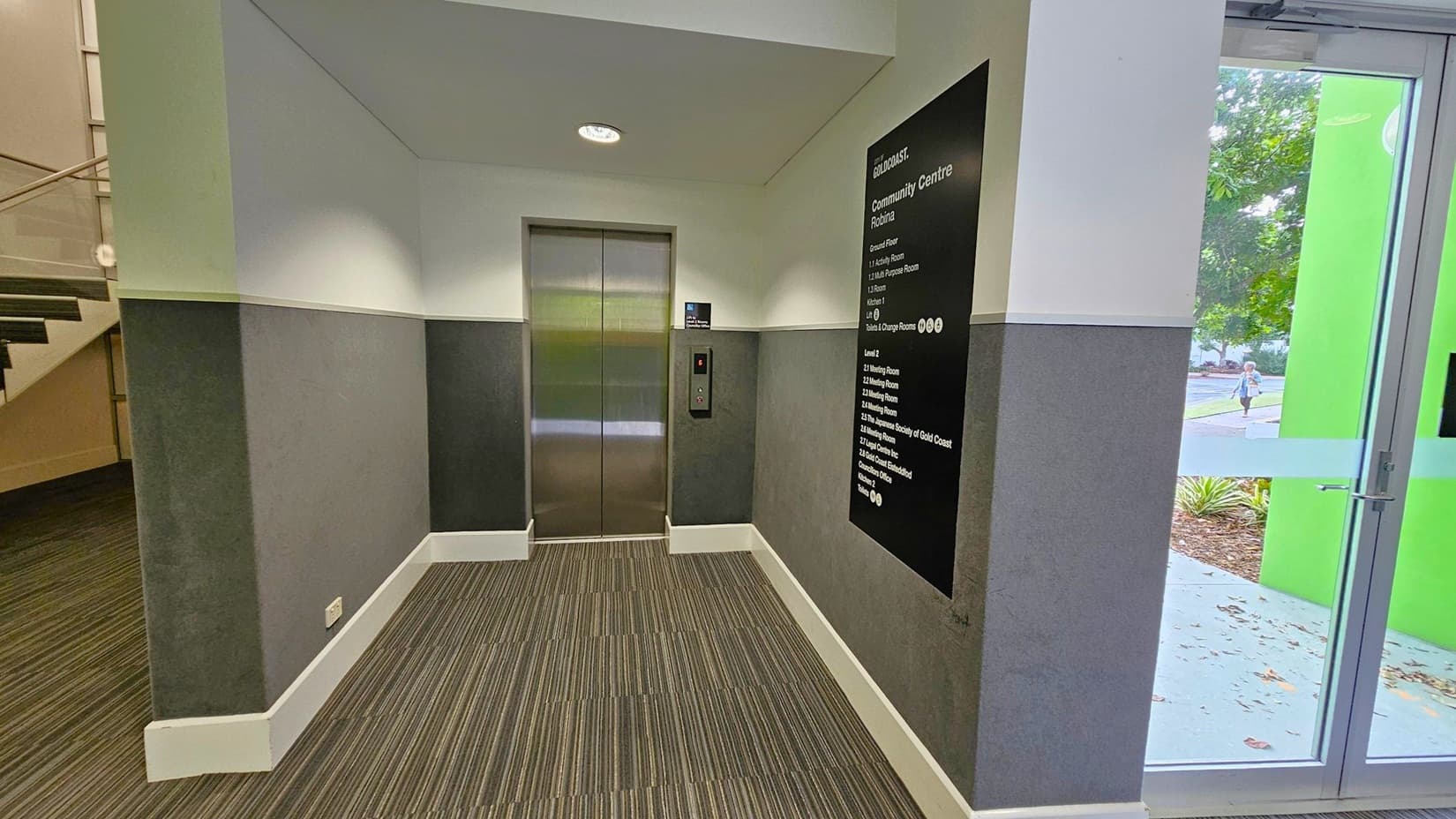 A hallway with a lift in the centre, featuring metallic doors. To the right, a black directory sign lists various rooms and facilities. A glass door with a green frame is also on the right, showing a view of the outside with trees and a person walking. On the left, a staircase with a wooden handrail is partially visible. The floor is covered with striped carpet, and the walls are painted in two tones of grey. The ceiling is plain with a recessed light.