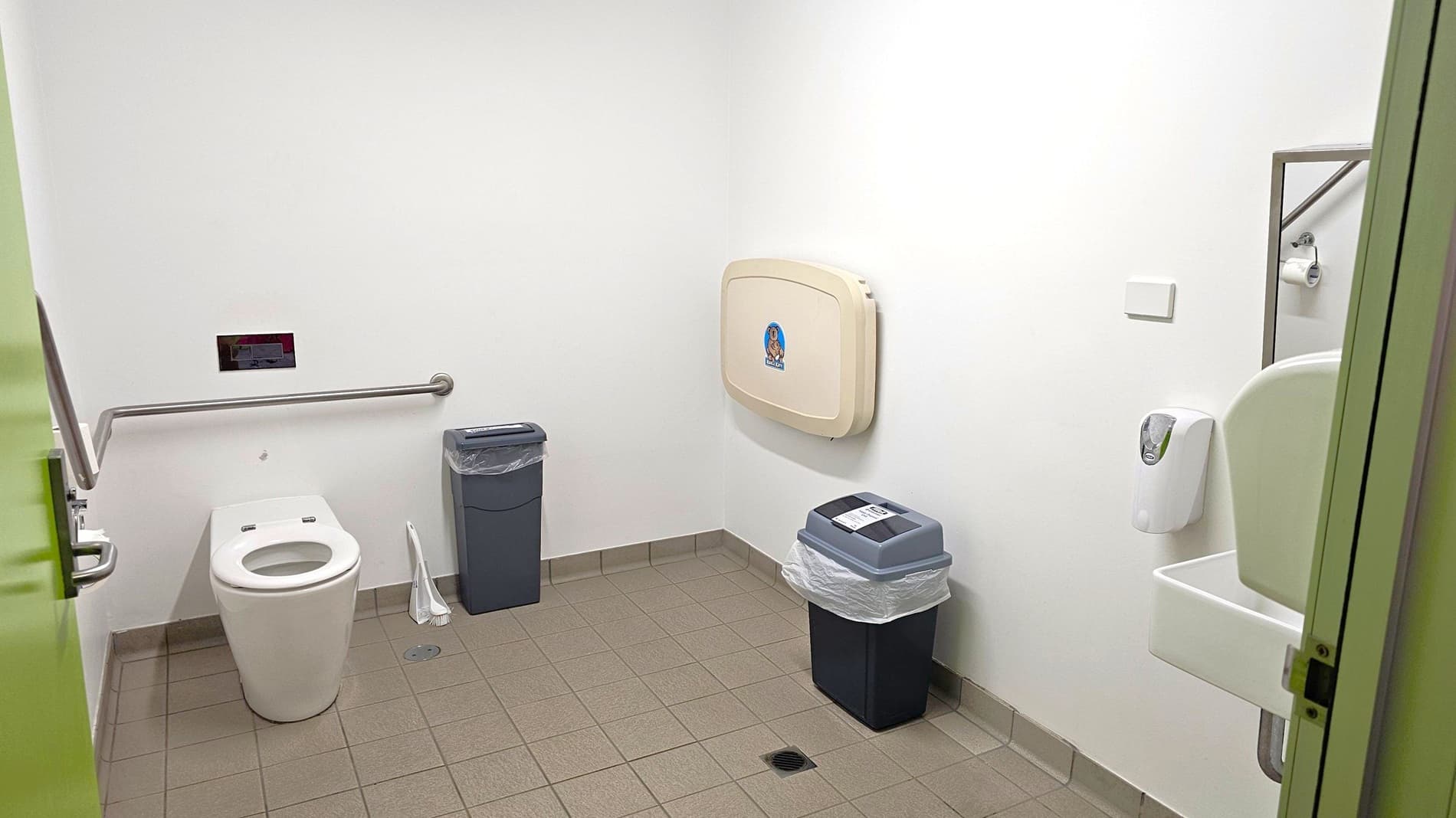 A public restroom with a white toilet on the left, equipped with a metal grab bar. Next to it is a grey bin and a toilet brush. On the right wall, a baby changing station is mounted above a black bin with a purple lid. A white soap dispenser is near a small sink, partially visible. The floor is tiled in light brown, and the walls are plain white. A green door is slightly open on the left.
