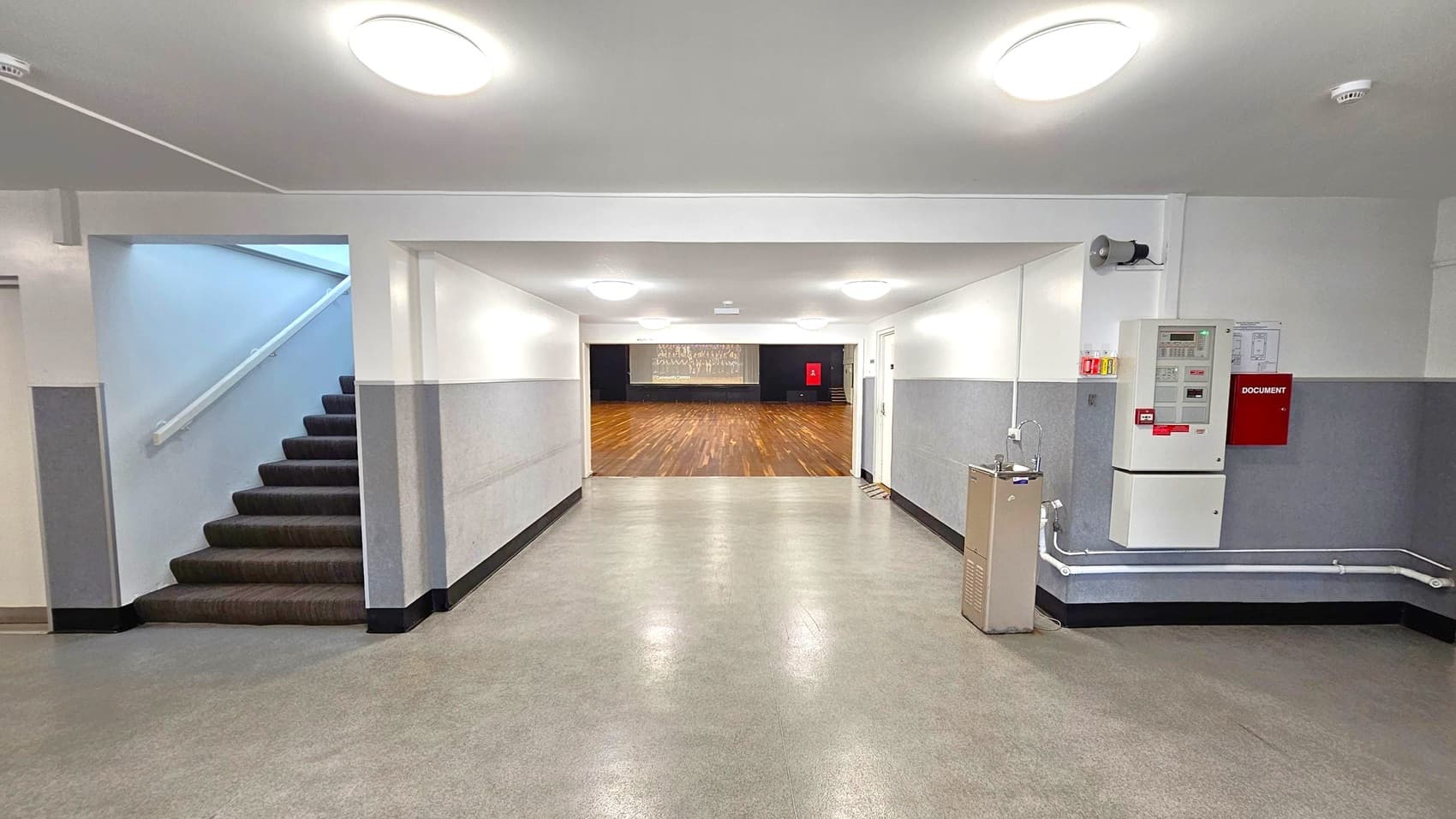 A hallway with a greenish floor and white ceiling featuring round lights. On the left, a staircase with dark steps leads upwards. On the right, a wall-mounted fire alarm system and a water fountain are visible. In the centre, the hallway opens into a large room with a wooden floor and a stage at the far end. The walls are grey with a white upper section.