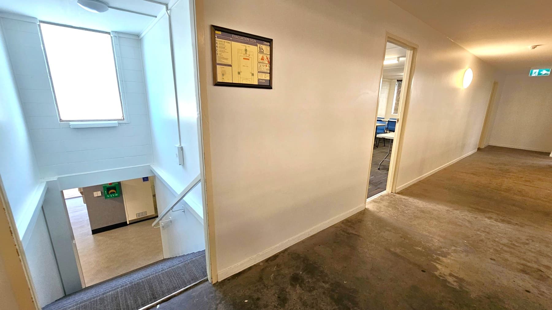 A hallway with a concrete floor and white walls. On the left, a staircase descends, with a window above allowing natural light. A framed evacuation sign is mounted on the wall near the stairs. On the right, an open door leads to a room with blue chairs and tables. Further down the hallway, an illuminated exit sign is visible. The ceiling is plain with a circular light fixture.