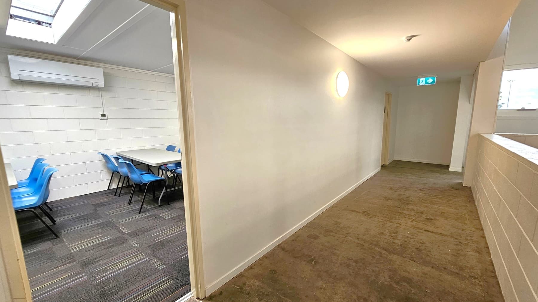 A hallway with a brown carpeted floor and white walls. On the left, an open door reveals a room with blue chairs around a table, a white brick wall, and an air conditioning unit. The ceiling has a skylight. The hallway has a round wall light and an exit sign above a door at the end. The right side features a half-wall with a window above it.