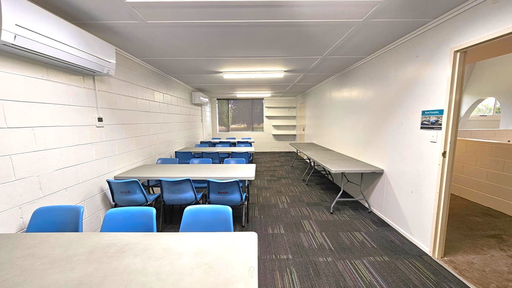 A classroom with white brick walls and a grey ceiling. On the left, there is an air conditioning unit mounted on the wall. Several tables with blue chairs are arranged in rows. On the right, a long table is against the wall, with shelves above it. The floor is covered with dark carpet tiles featuring subtle coloured lines. At the back, a window with a blind is visible. A doorway on the right leads to another area, with a sign on the wall next to it.