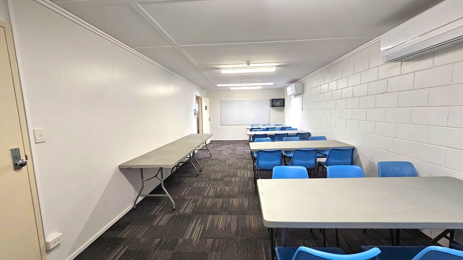 A room with white walls and a patterned carpeted floor. On the left, a long folding table is positioned against the wall. On the right, several tables with blue plastic chairs are arranged in rows. The back wall features a whiteboard and a wall-mounted television. The ceiling has fluorescent lights and an air conditioning unit is mounted on the right wall. A door is visible at the far end of the room.