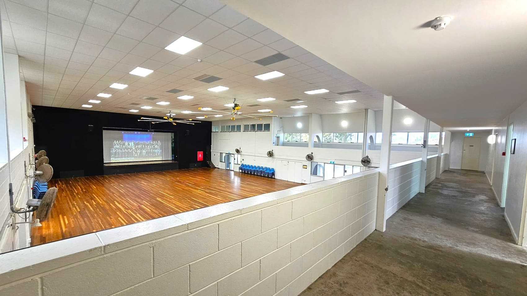 A large indoor hall with a wooden floor and a stage at the far end displaying a projected image. The ceiling is fitted with numerous square lights and ceiling fans. On the left, there are several wall-mounted fans and stacked blue chairs. The right side features a corridor with a carpeted floor and a railing overlooking the hall. The walls are light-coloured, and there are windows along the upper part of the walls.