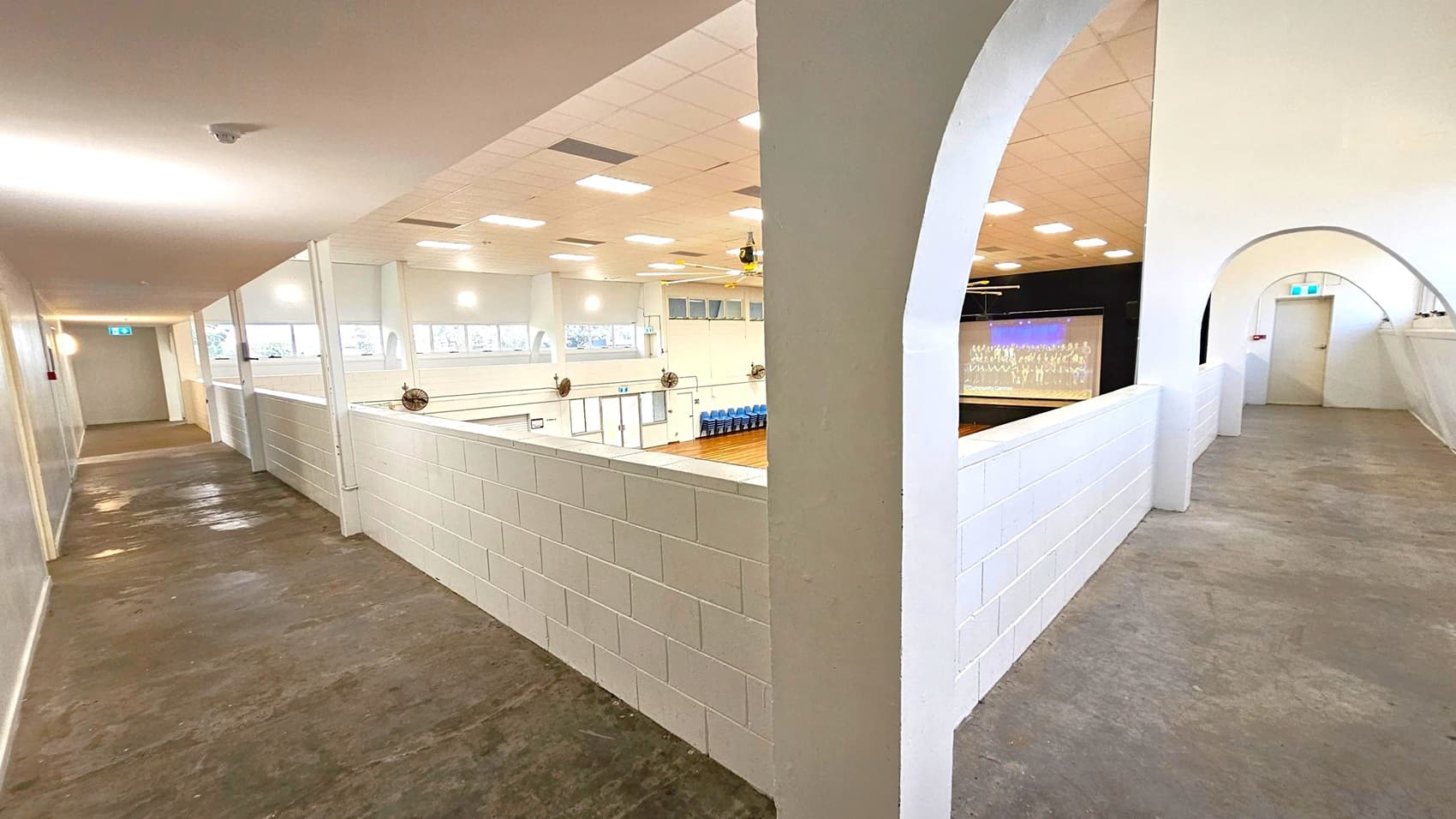 A corridor with a concrete floor and white ceiling. On the left, a long hallway with windows and a door at the end. In the middle, a low white brick wall overlooks a gymnasium or auditorium with wooden flooring and blue chairs. On the right, an arched walkway with a door at the end. Ceiling lights are evenly spaced, and a projector screen is visible in the auditorium area.