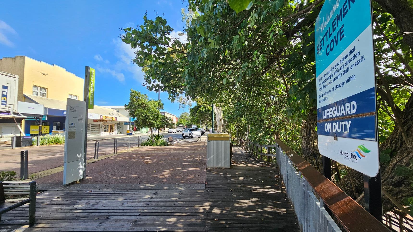A wooden boardwalk runs along the right side, bordered by dense green foliage and a sign reading "Settlement Cove" with "Lifeguard on Duty." On the left, a paved walkway leads past a row of shops with colourful signage. A bench and a bin are visible on the boardwalk. In the background, a street with parked cars and more trees can be seen under a clear blue sky.