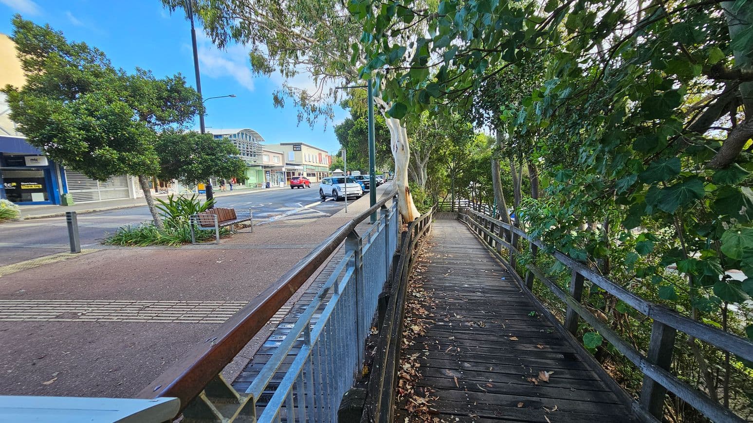A wooden walkway on the right is bordered by lush green trees and a metal railing. On the left, a street with parked cars and a row of shops, including a blue storefront, is visible. A bench and a small garden with a tree are situated near the pavement. The ground is a mix of concrete and reddish paving, and a tall streetlamp is present. The scene is urban with a touch of greenery.