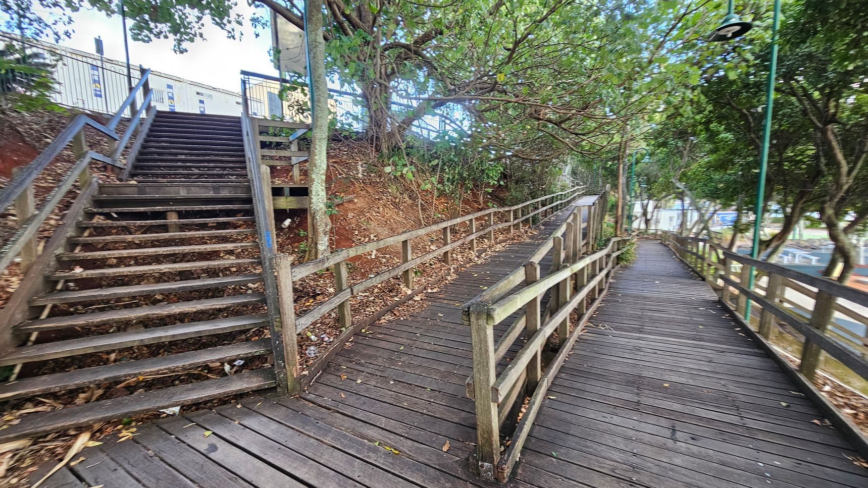 A wooden walkway with railings runs along the right side, bordered by trees and foliage. On the left, a set of wooden stairs ascends a small hill, also surrounded by trees. The ground is covered with fallen leaves. In the background, a building with a metal fence is visible at the top of the stairs. A street lamp is positioned along the walkway.