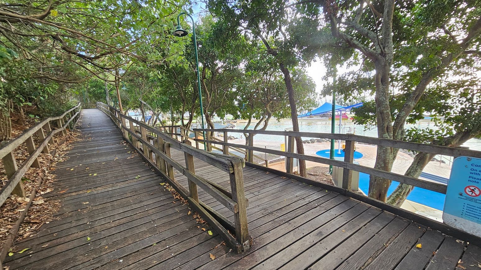 A wooden boardwalk curves to the left, bordered by a wooden railing. On the left, dense foliage with overhanging branches creates a shaded path. On the right, trees line the path, and beyond them, a pool area with a blue shade structure is visible. The ground is scattered with dry leaves, and a lamp post stands along the path. A sign with text is partially visible on the right.