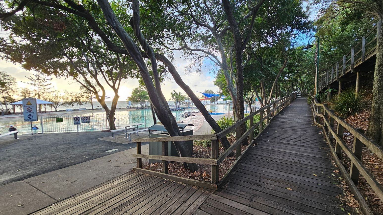 A wooden boardwalk runs along the right side, bordered by a wooden railing and surrounded by dense trees and foliage. On the left, there's a fenced swimming pool area with a few people in the water. Signs are visible near the pool, and there are benches and a bin nearby. The ground is a mix of concrete and wooden planks, with scattered leaves. The background features more trees and a glimpse of the sky.