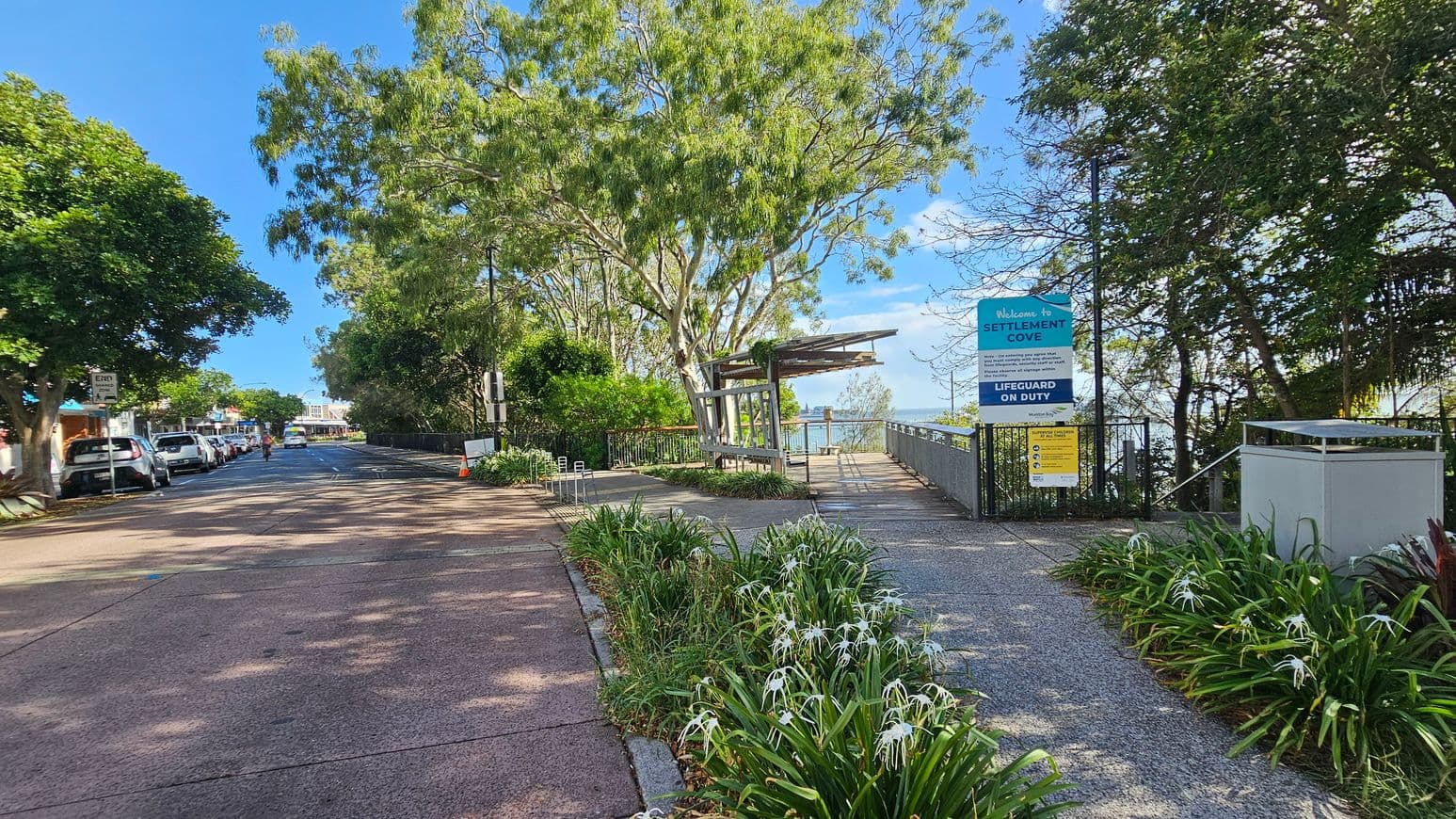 A pathway leads to a waterfront area on the right, with a sign reading "Welcome to Settlement Cove" and "Lifeguard on Duty". The path is lined with green plants and white flowers. On the left, a street with parked cars and a pedestrian is visible, bordered by trees. A large tree provides shade over the path, and a wooden structure is near the water's edge. The sky is clear and blue.